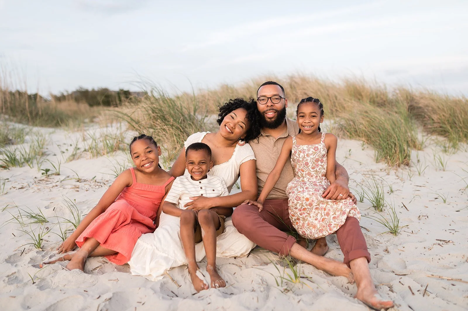 A happy family of six sitting on the beach with sand dunes and grass behind them, smiling for the camera.