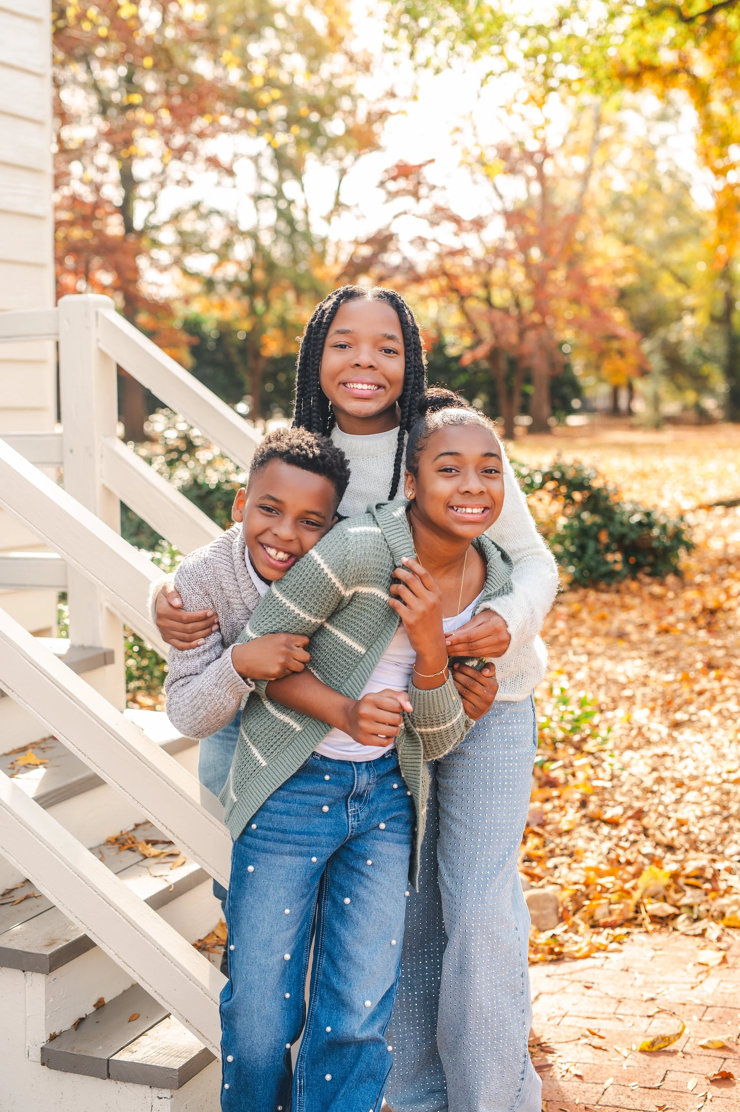 Three children smiling and hugging on stairs outdoors during fall, with orange and yellow leaves on trees and ground.
