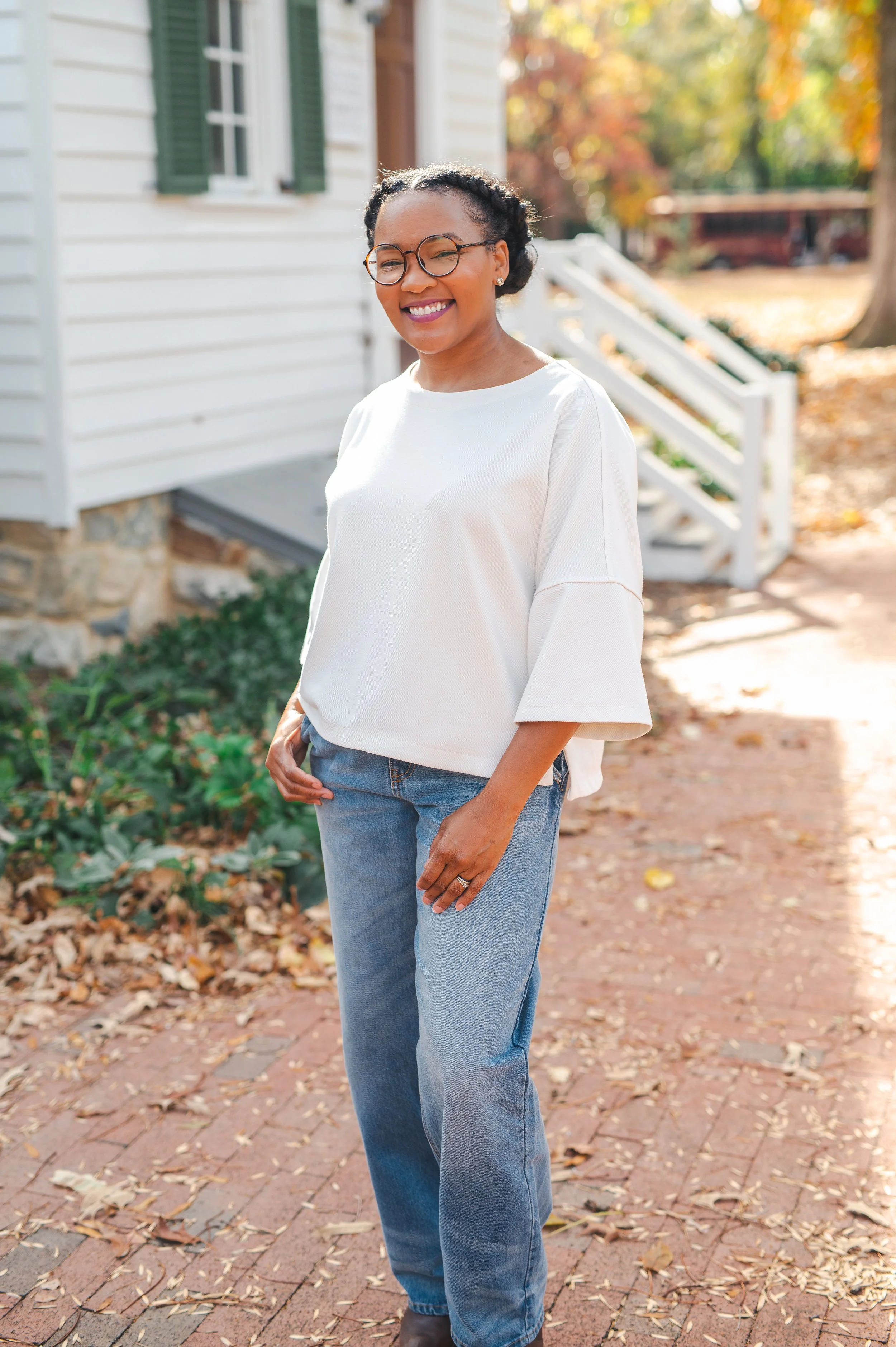 A woman smiling outdoors on a brick pathway, wearing glasses, a loose white top with three-fourths sleeves, and blue jeans, with autumn leaves on the ground and a white house with green shutters in the background.