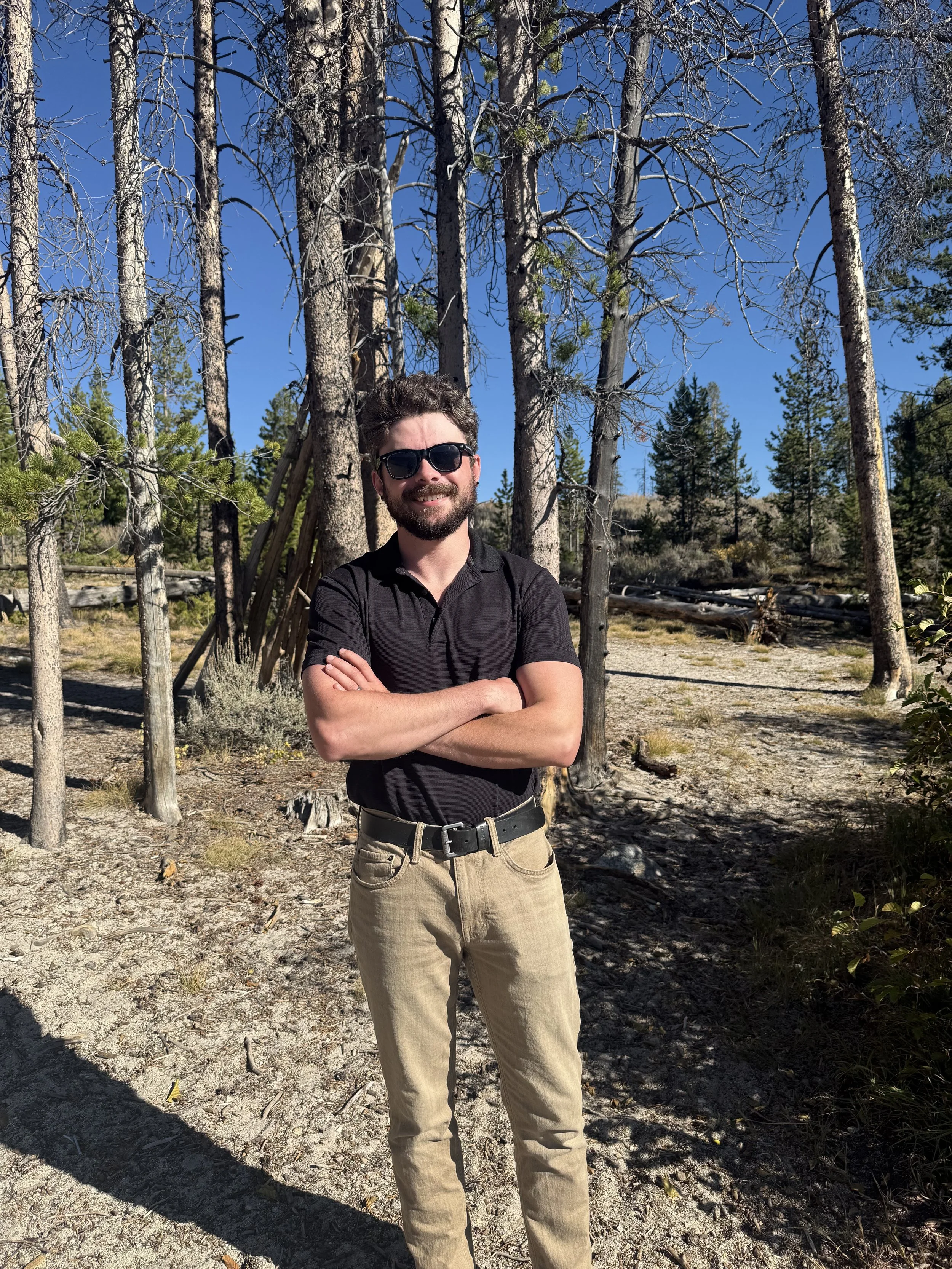 A man with dark hair and a beard, wearing sunglasses, a black polo shirt, and khaki pants, stands with his arms crossed in a forest with tall trees and a clear blue sky.