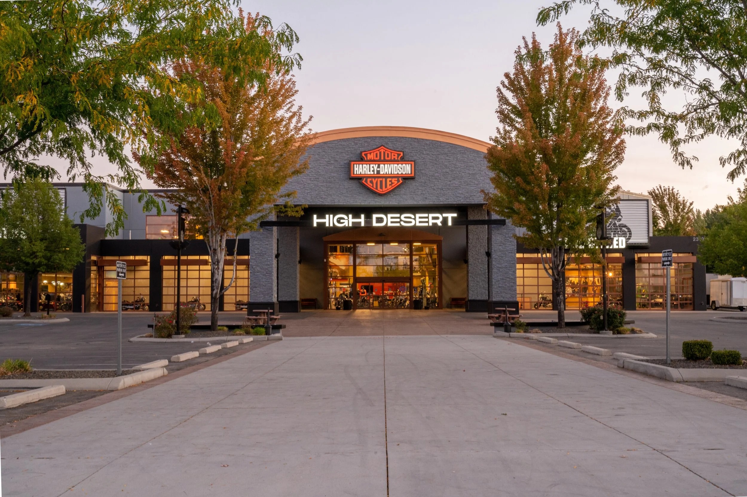 Exterior view of a Harley-Davidson motorcycle dealership called High Desert, with trees and parking lot in front, illuminated signage, and large windows showing the inside display area.