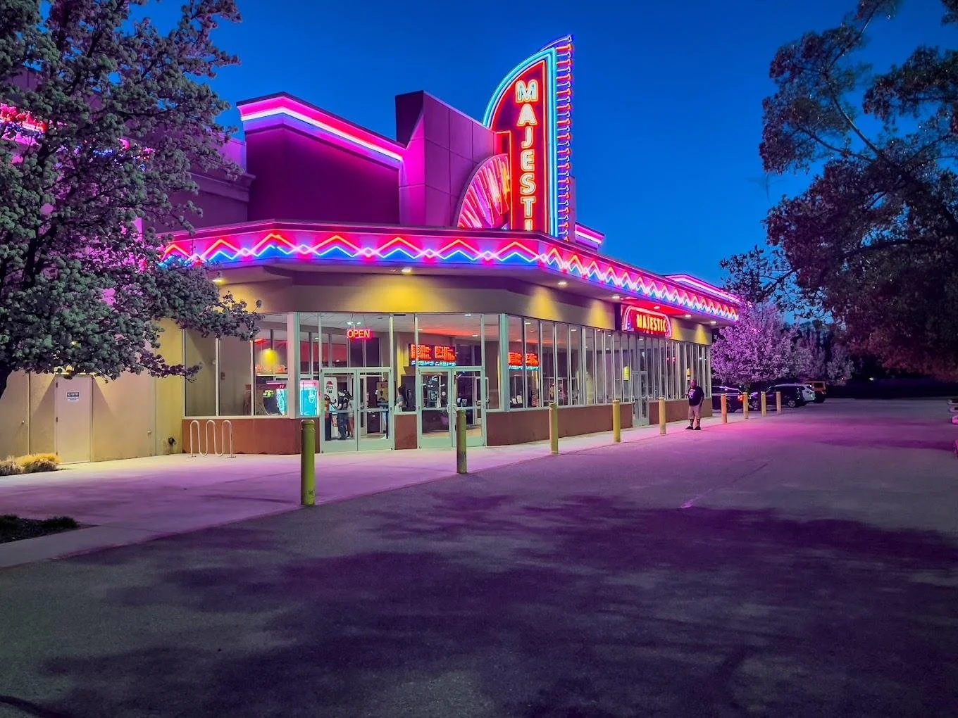 Nighttime view of a vintage-style diner called 'Majestic' with bright neon pink, purple, and blue lights, glass entrance doors, surrounded by trees and a parking lot with a few cars.