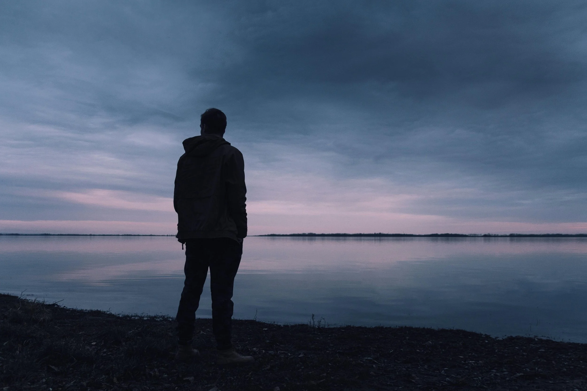 A person standing alone on a dark shoreline, looking out over a calm lake with a cloudy sky overhead at dusk or dawn.