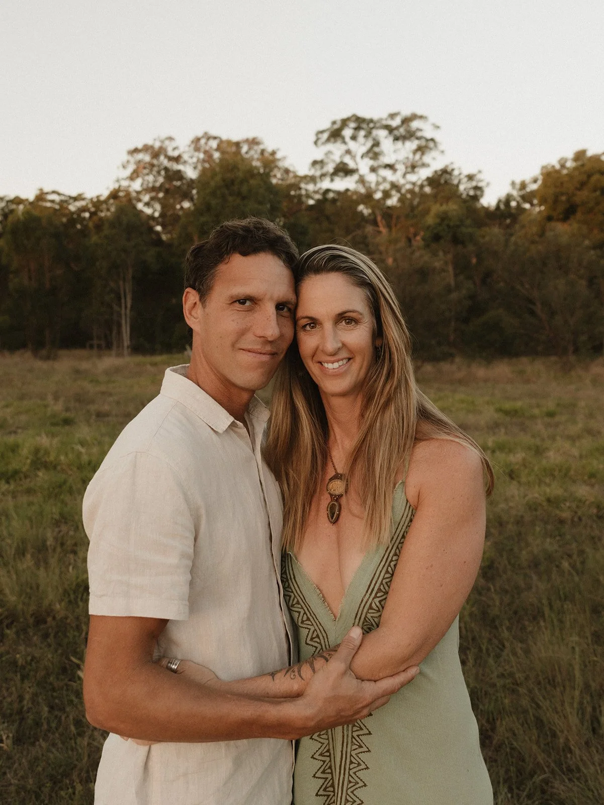 A couple standing close together outdoors during sunset, with trees in the background. The man has short dark hair and wears a light-colored linen shirt. The woman has long, wavy hair and wears a sleeveless green dress with embroidery, and a pendant necklace. They are smiling and the man is holding her arm.