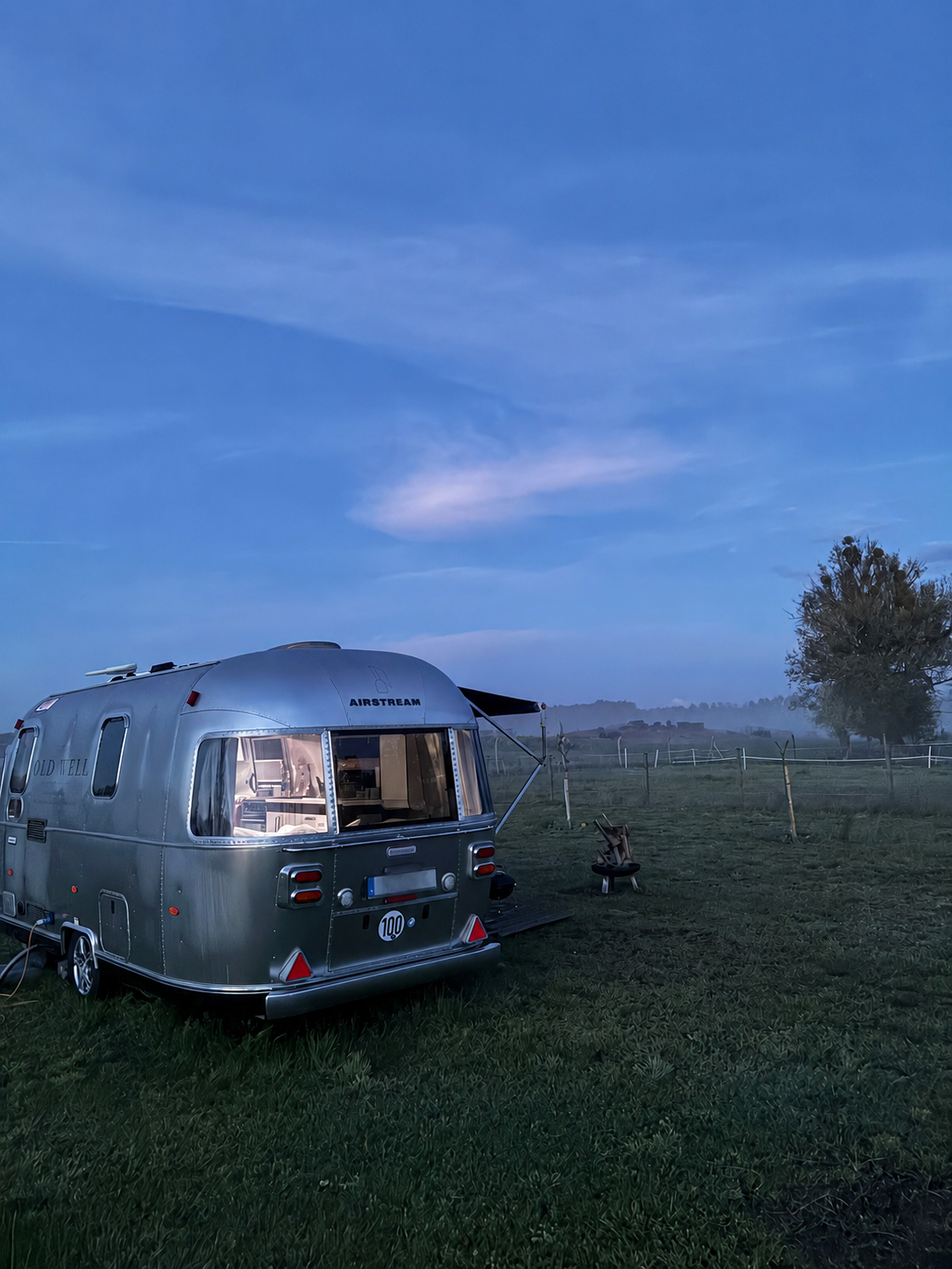 A silver Airstream travel trailer parked on a grassy field at dusk, with a tree and a fence in the background under a blue sky with scattered clouds.