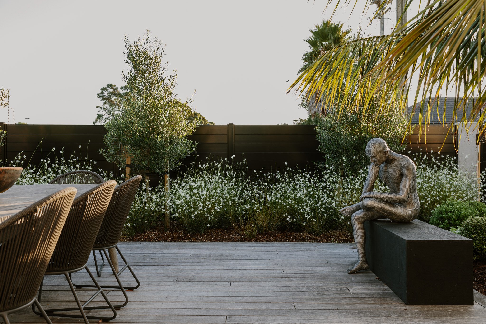 A modern outdoor patio with wooden decking, a long dining table with wicker chairs on the left, a black bench on which a metallic sculpture of a seated woman is placed, green plants, and white flowering shrubs against a dark fence, with palm trees an