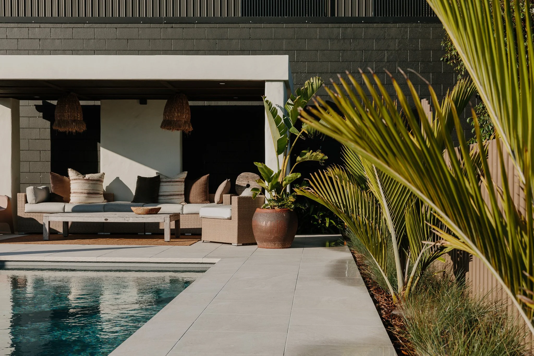 Outdoor patio with a swimming pool, beige wicker sofa with pillows, potted plants, and tropical foliage.