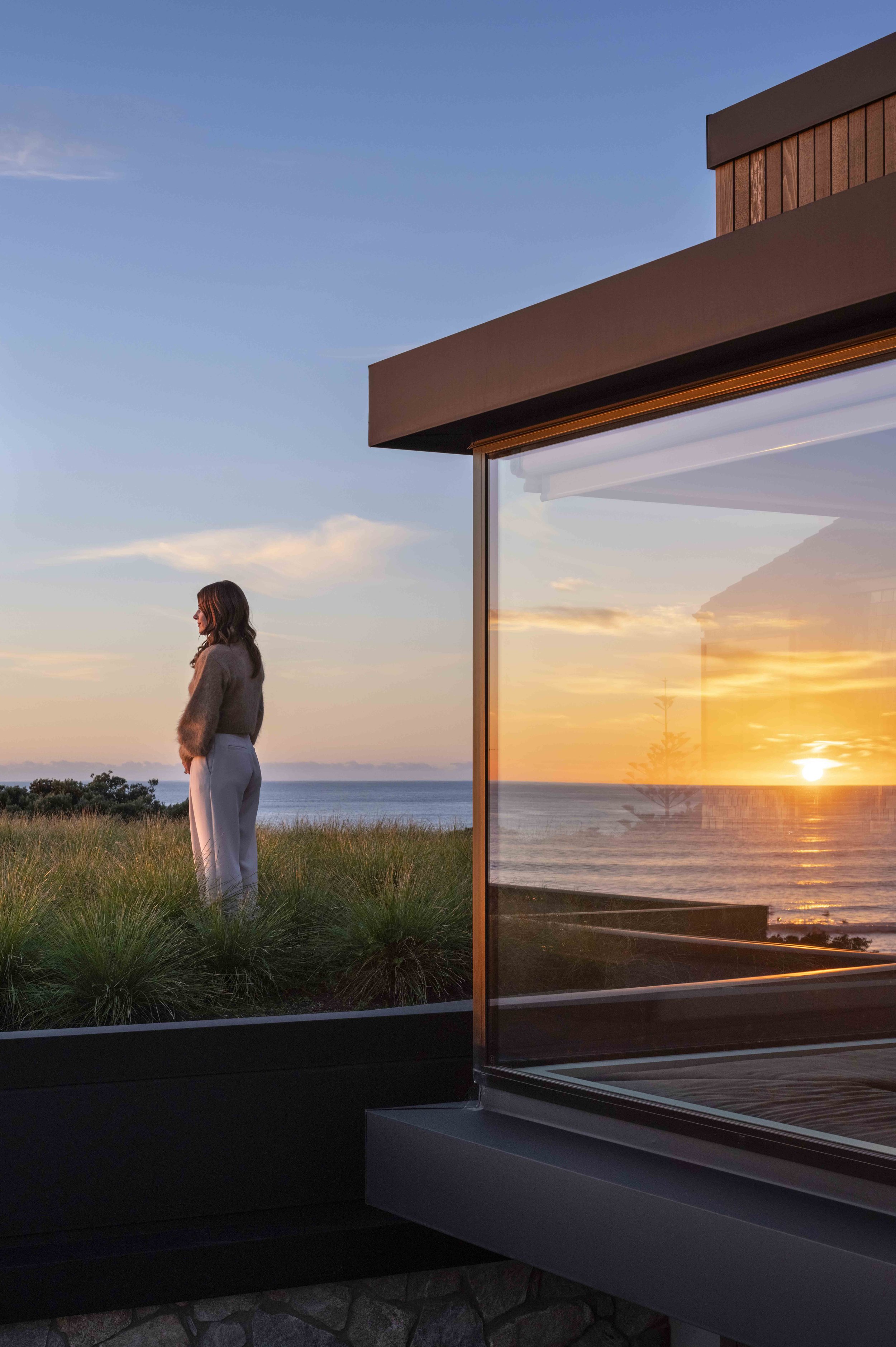 A woman with brown hair standing on a grassy rooftop garden, looking at the sunset over the ocean, with a modern house reflecting the sunset in large glass windows.
