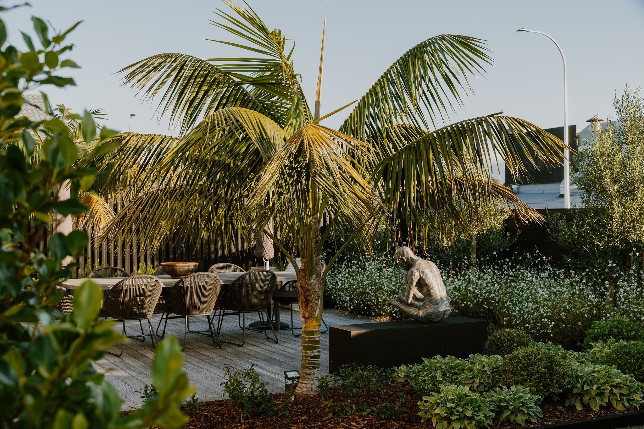 A patio with a large palm tree, outdoor dining table with chairs, and a sculpture of a person sitting, surrounded by plants and flowers, with city buildings in the background.