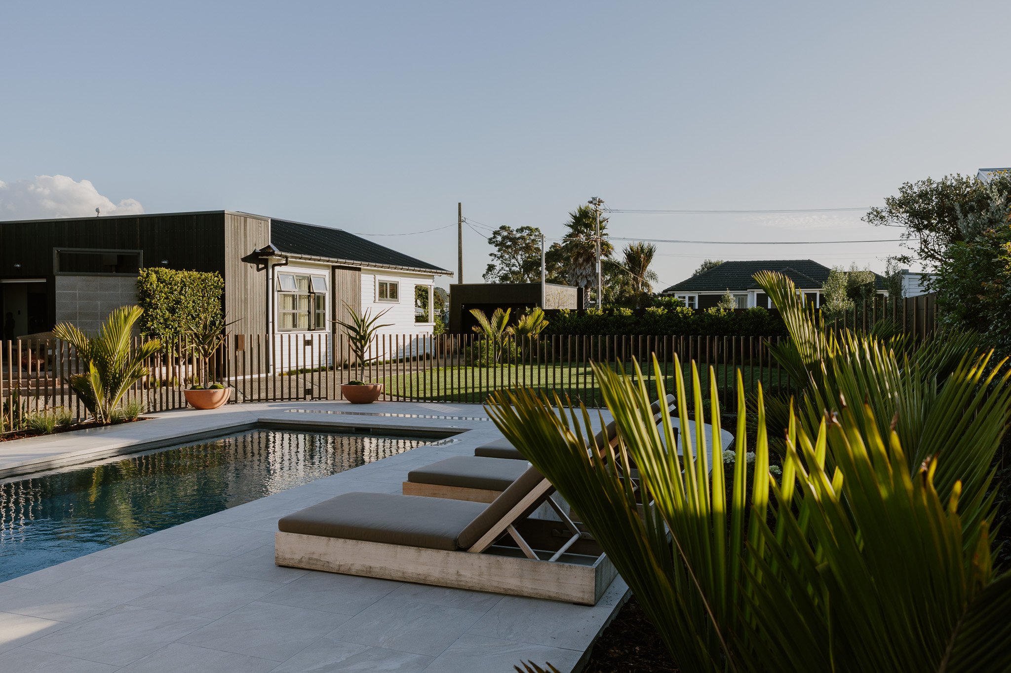 backyard with pool, lounge chairs, potted plants, wooden fence, green lawn, and houses in the background under a clear sky