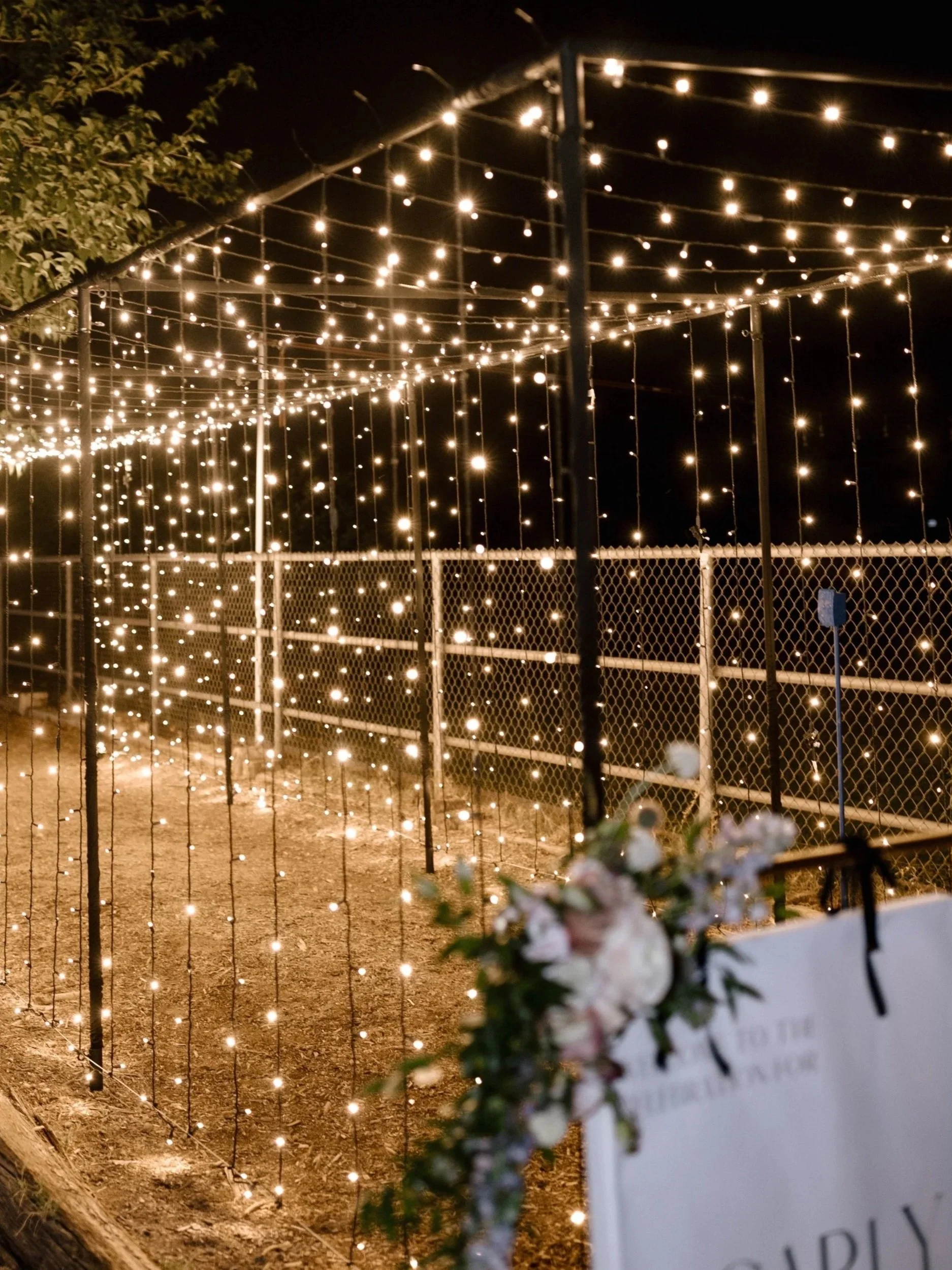 String of glowing string lights hanging above a chain-link fence at night, with a blurred sign and flowers in the foreground.