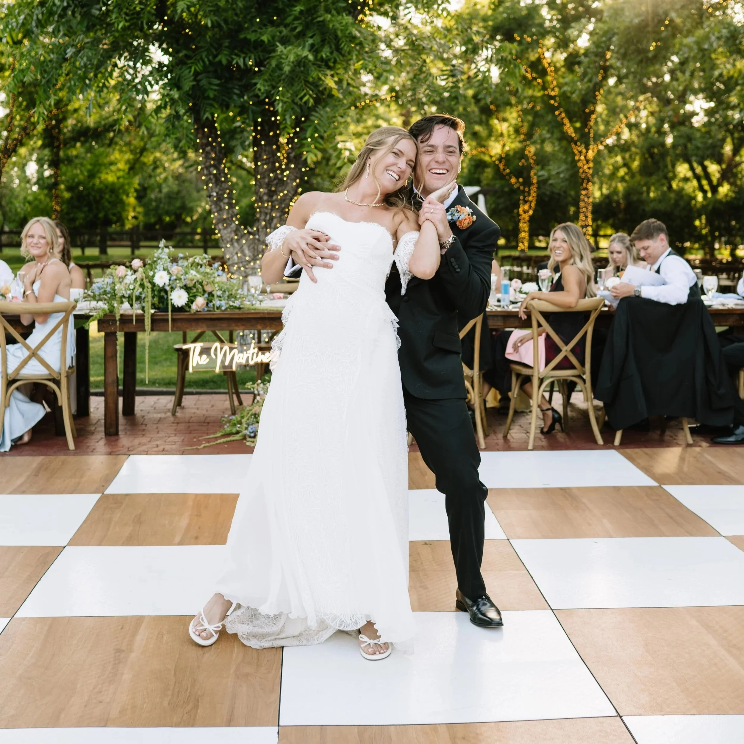 A bride and groom dancing at their wedding reception outdoors, with guests seated at tables in the background and string lights hanging from trees.