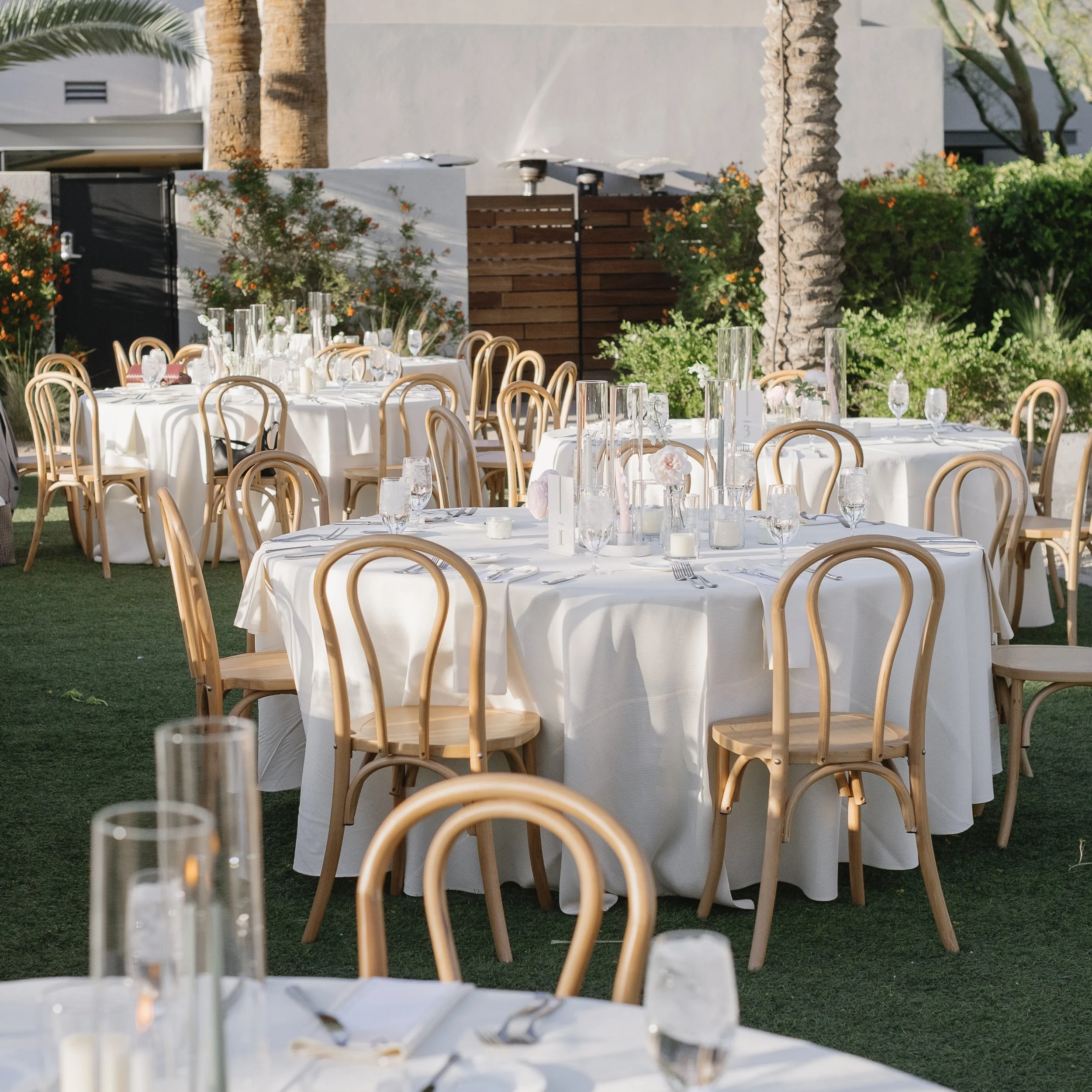 Outdoor wedding or event reception with round tables covered in white tablecloths, decorated with glassware, candles, and centerpieces, set on a grassy area with trees and bushes in the background.