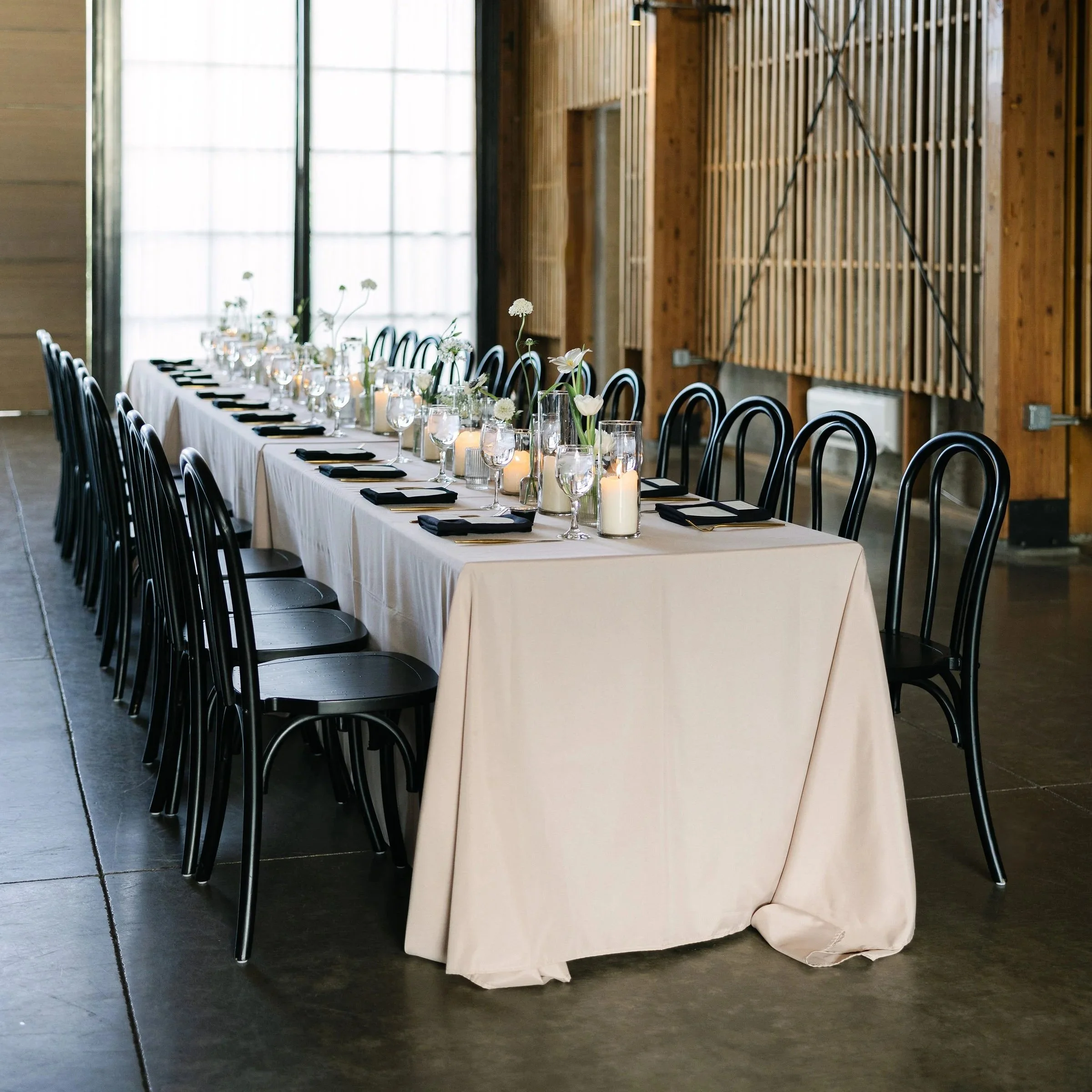 Elegant wedding or event reception table set with black chairs, white tablecloth, flower centerpieces, candles, glasses, and place settings in a modern venue with wood and metal accents.