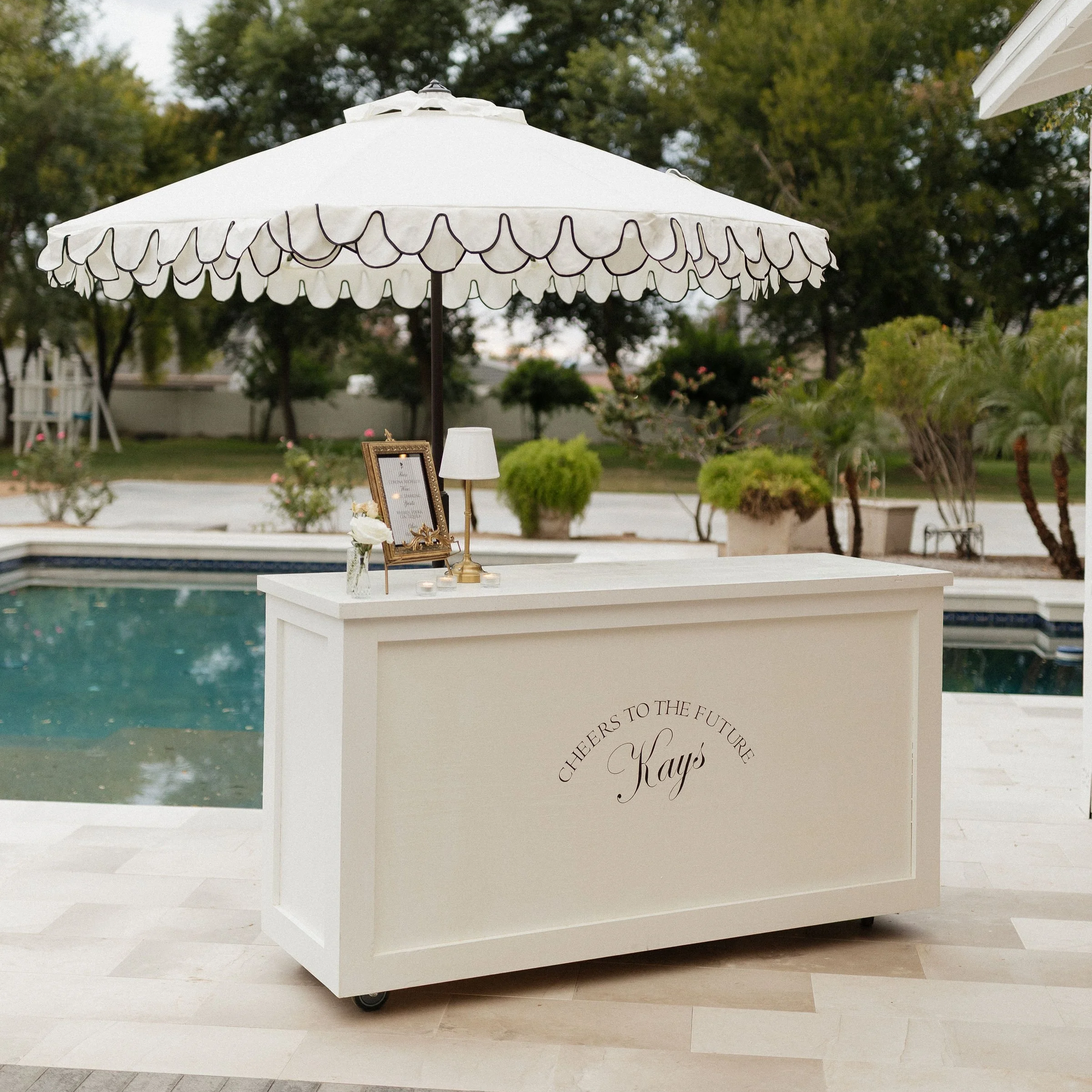 A white reception or guest sign-in desk with custom text 'Cheers to the future, Kays' on the front, located outdoors by a pool under a large white parasol with a scalloped edge. The desk holds a framed sign, a small lamp, and a vase of flowers. There
