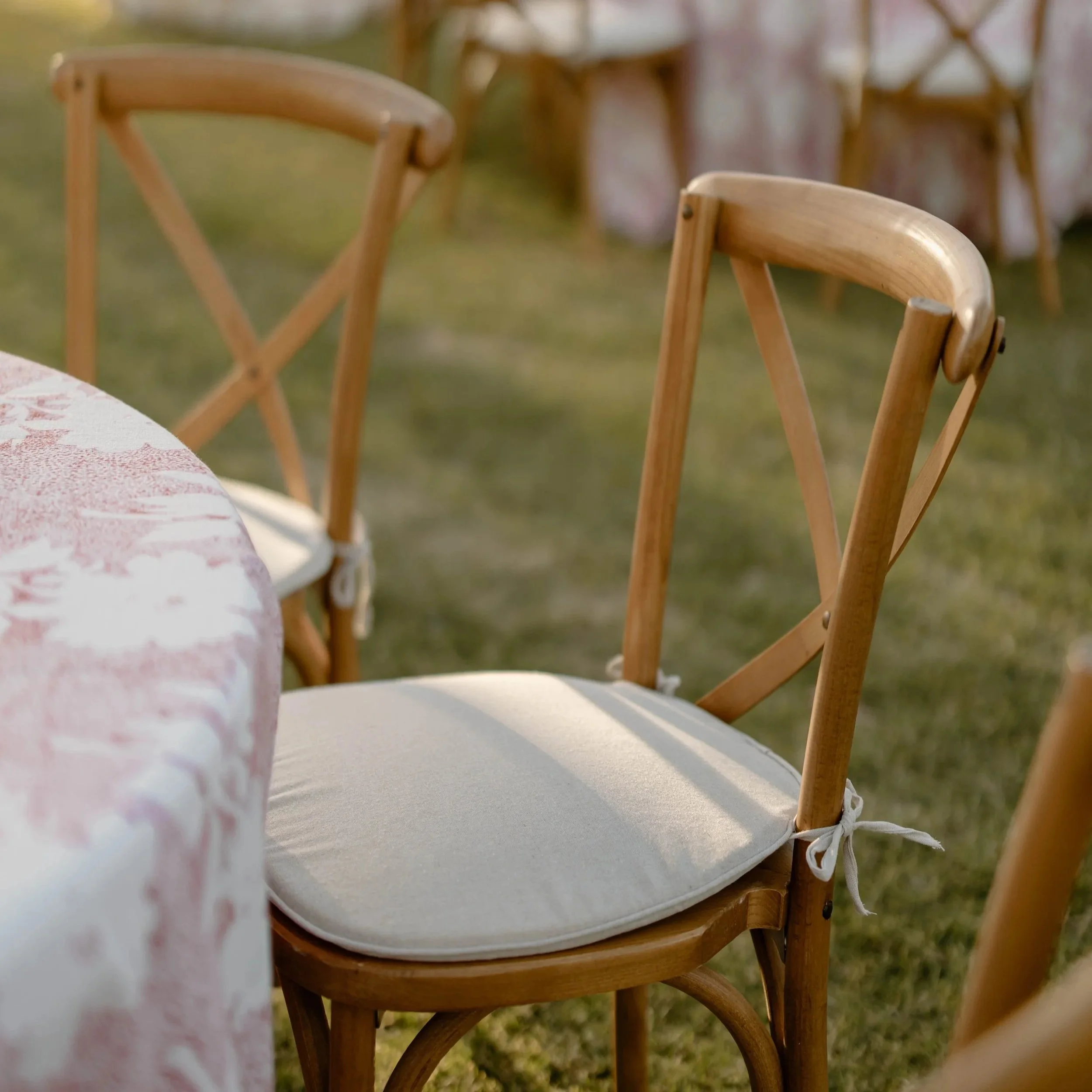 Wooden chairs with white cushions around a table with a pink and white tablecloth, outdoors on grass.