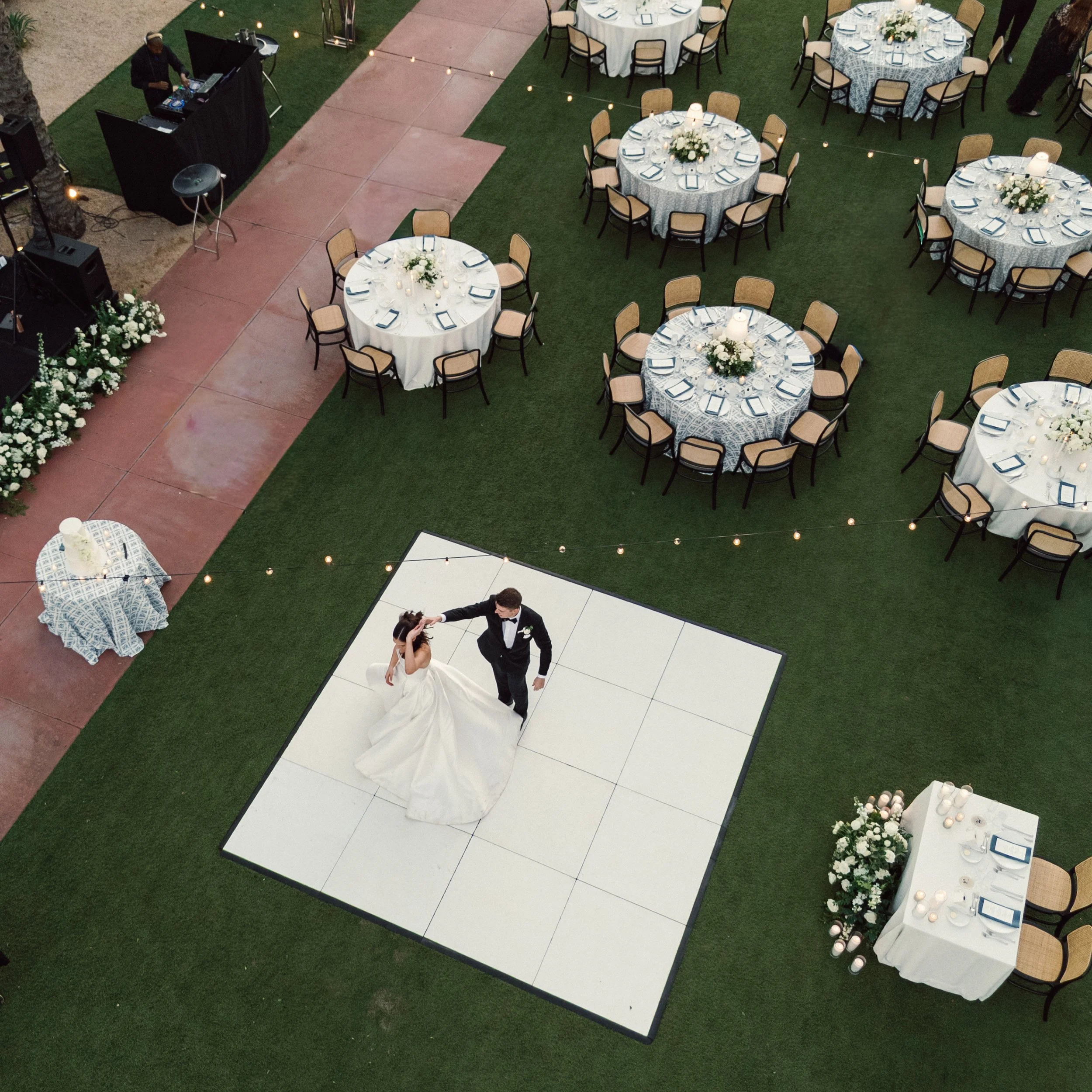 A bride and groom dancing on a white dance floor at an outdoor wedding reception, surrounded by round tables with white tablecloths and floral centerpieces, on a grass lawn with string lights overhead.