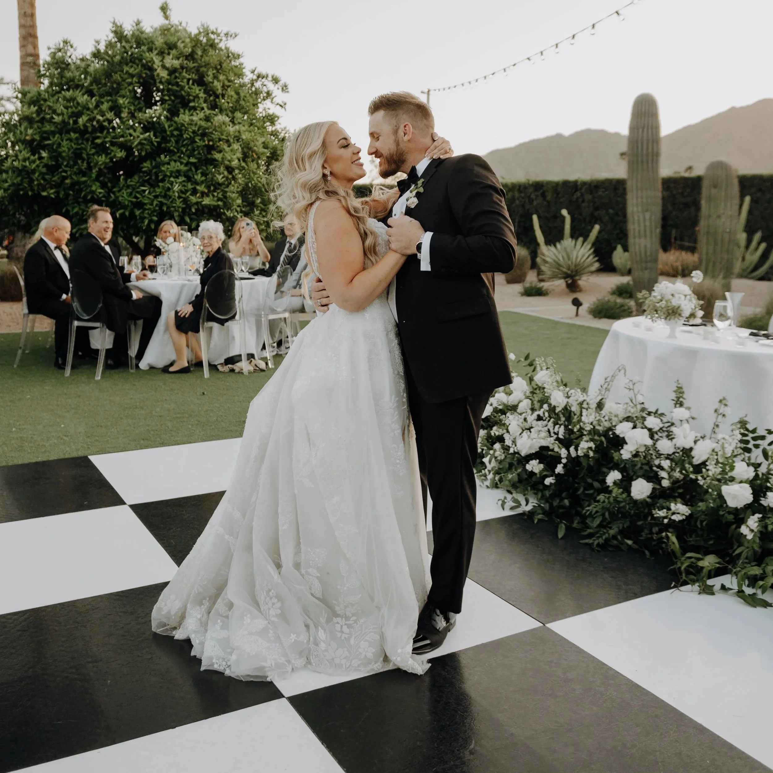 Bride and groom dancing at their wedding reception outdoors, with guests seated at tables in the background, in a desert landscape with cacti and mountains.