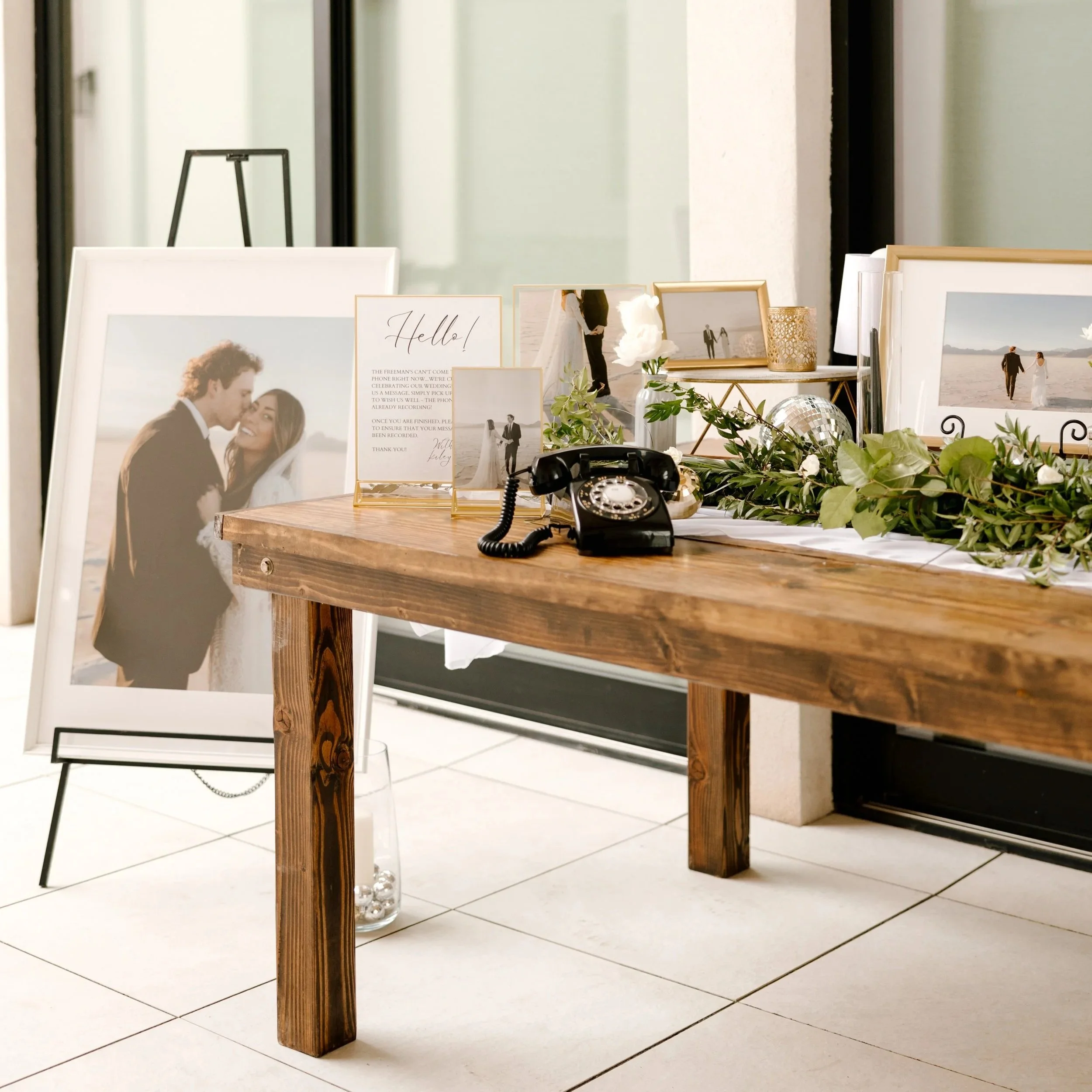 Display table with wedding photo frames, a vintage black rotary phone, and decorative green foliage.