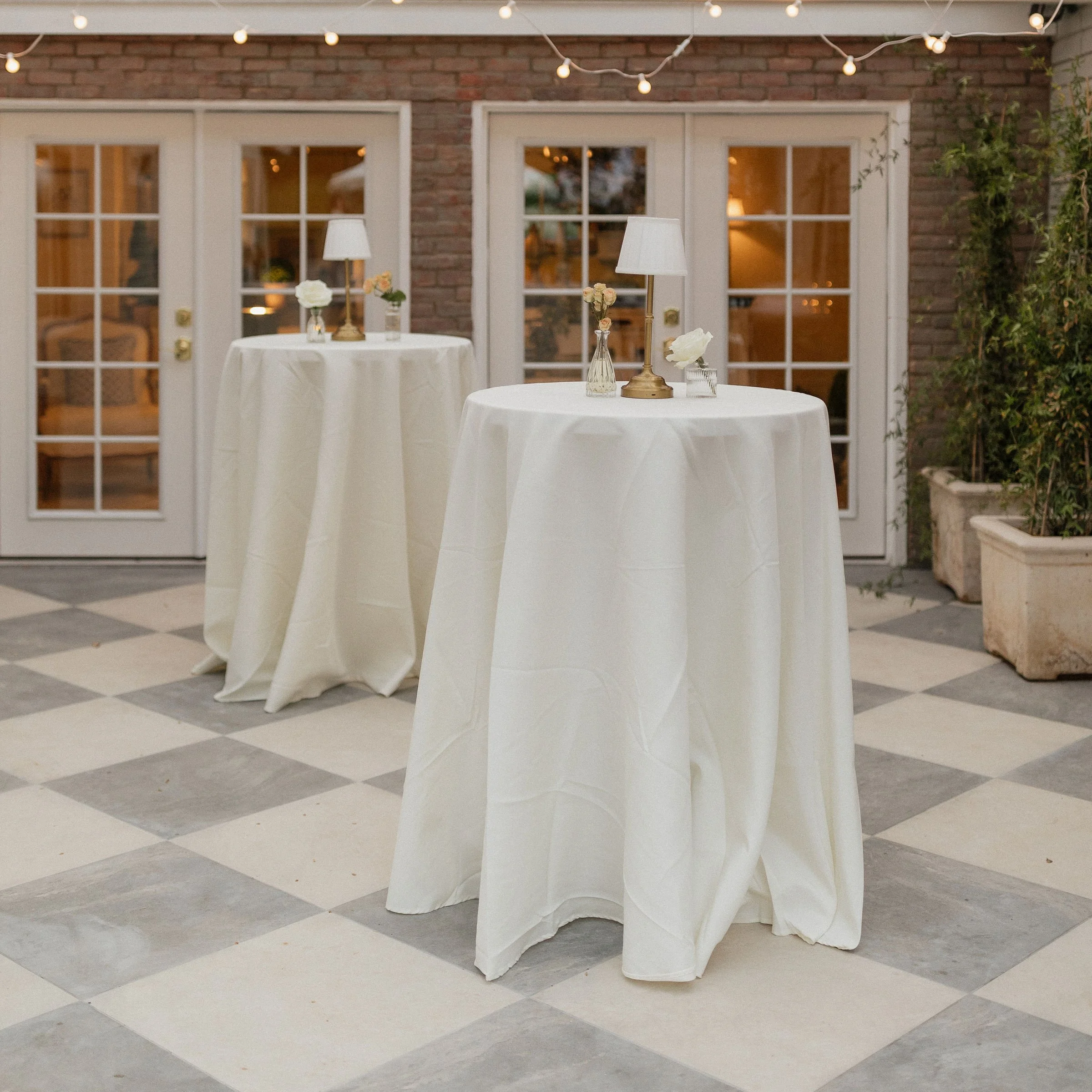 A small round table covered with a beige tablecloth outdoors, with a single white flower in a small glass vase on top. In the background, there are bushes, trees, and a white building.