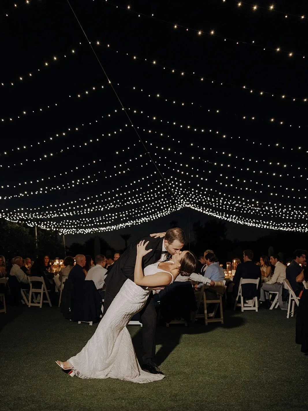 Outdoor evening wedding reception with round tables, white chairs, and string lights overhead, surrounded by trees and tall palm trees, under a dark sky.