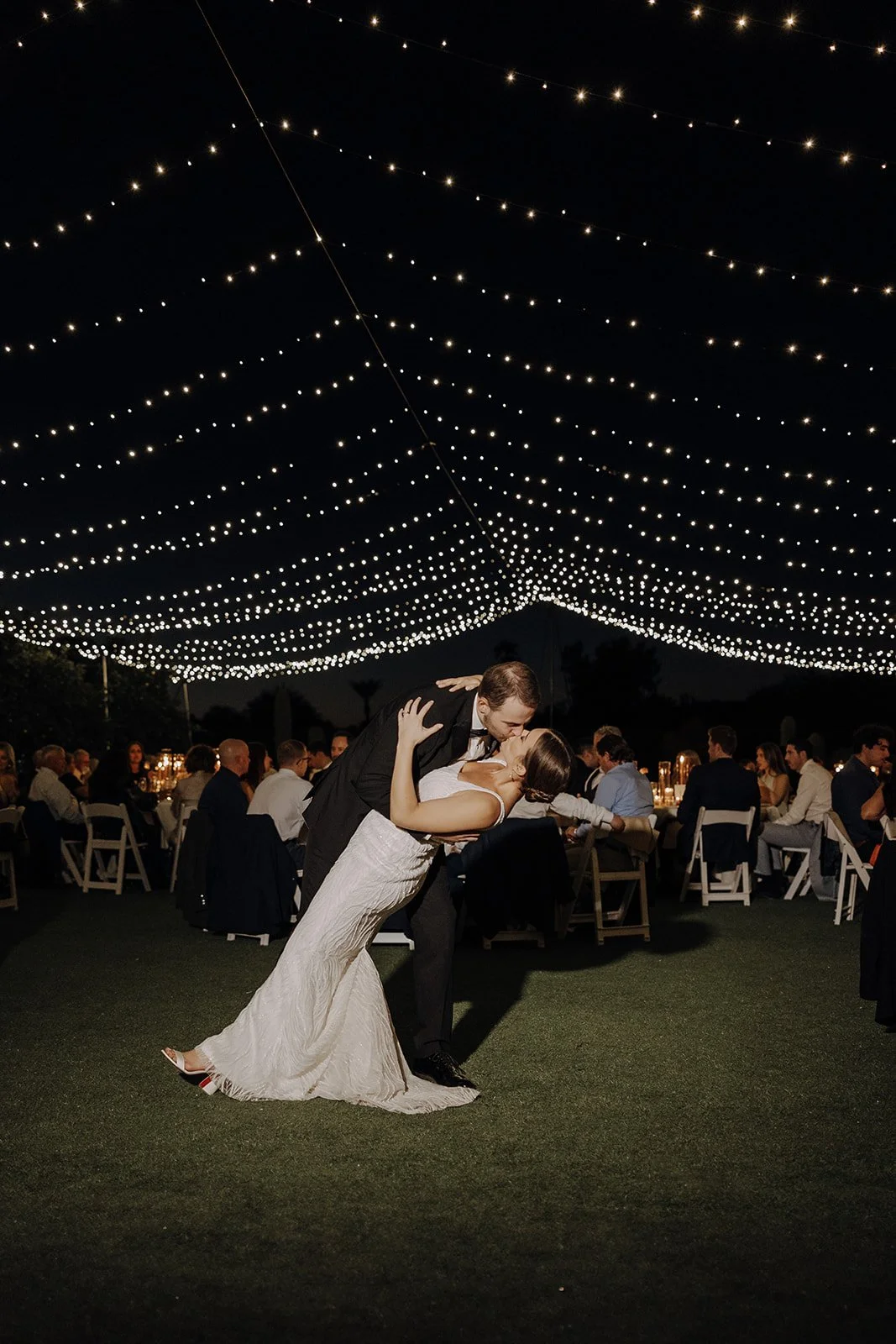 Outdoor evening wedding reception with round tables, white chairs, and string lights overhead, surrounded by trees and tall palm trees, under a dark sky.