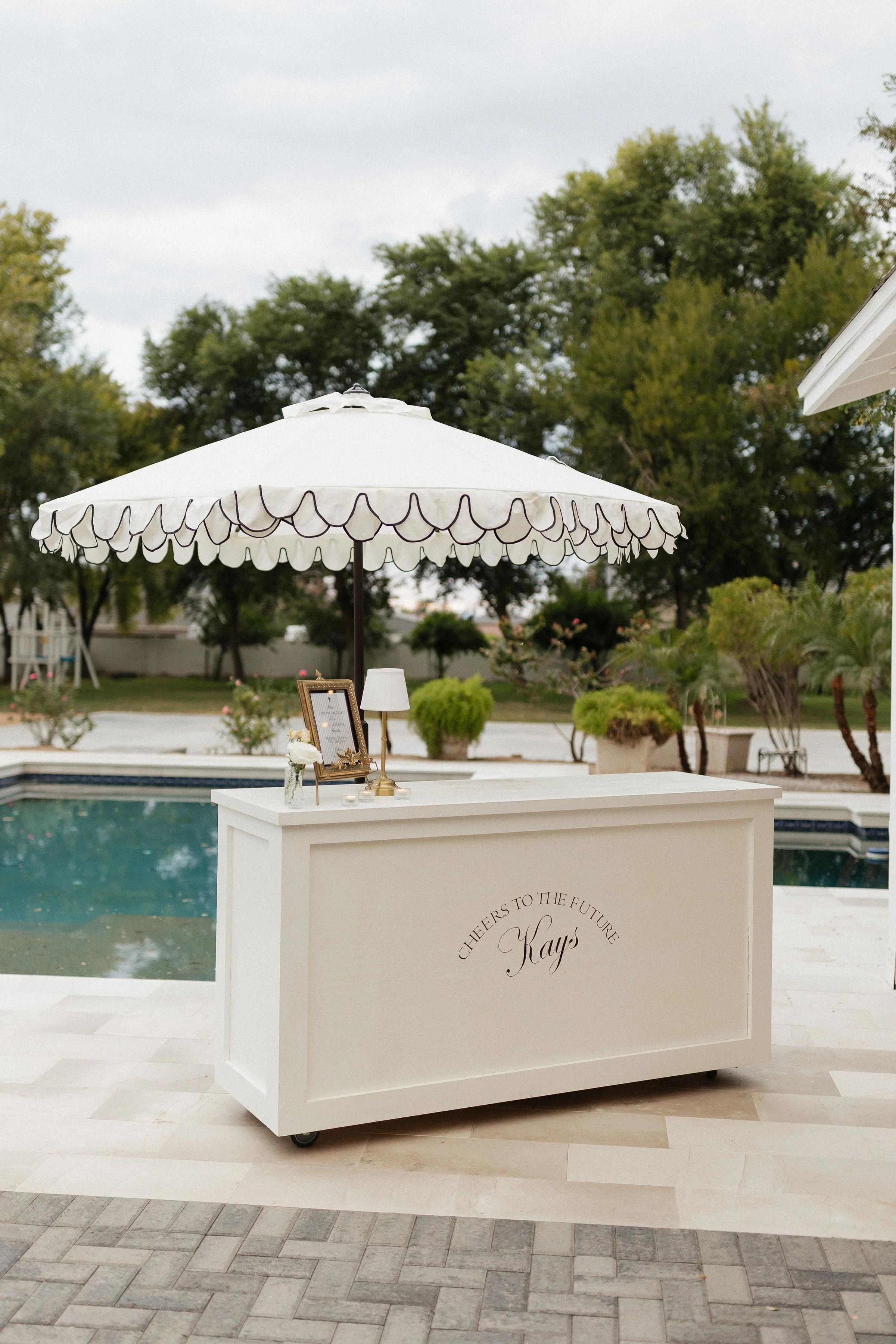 A white drinks or concession stand with an umbrella, set outdoors near a swimming pool and surrounded by plants and trees.