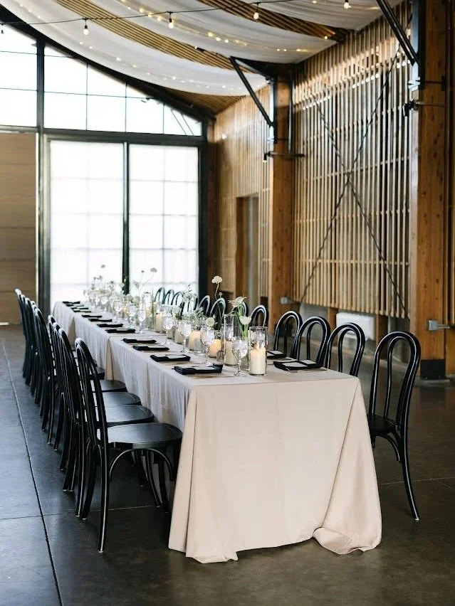 Interior of a modern, rustic event space with a long banquet table set for a meal, decorated with candles and flowers, and surrounded by black chairs.