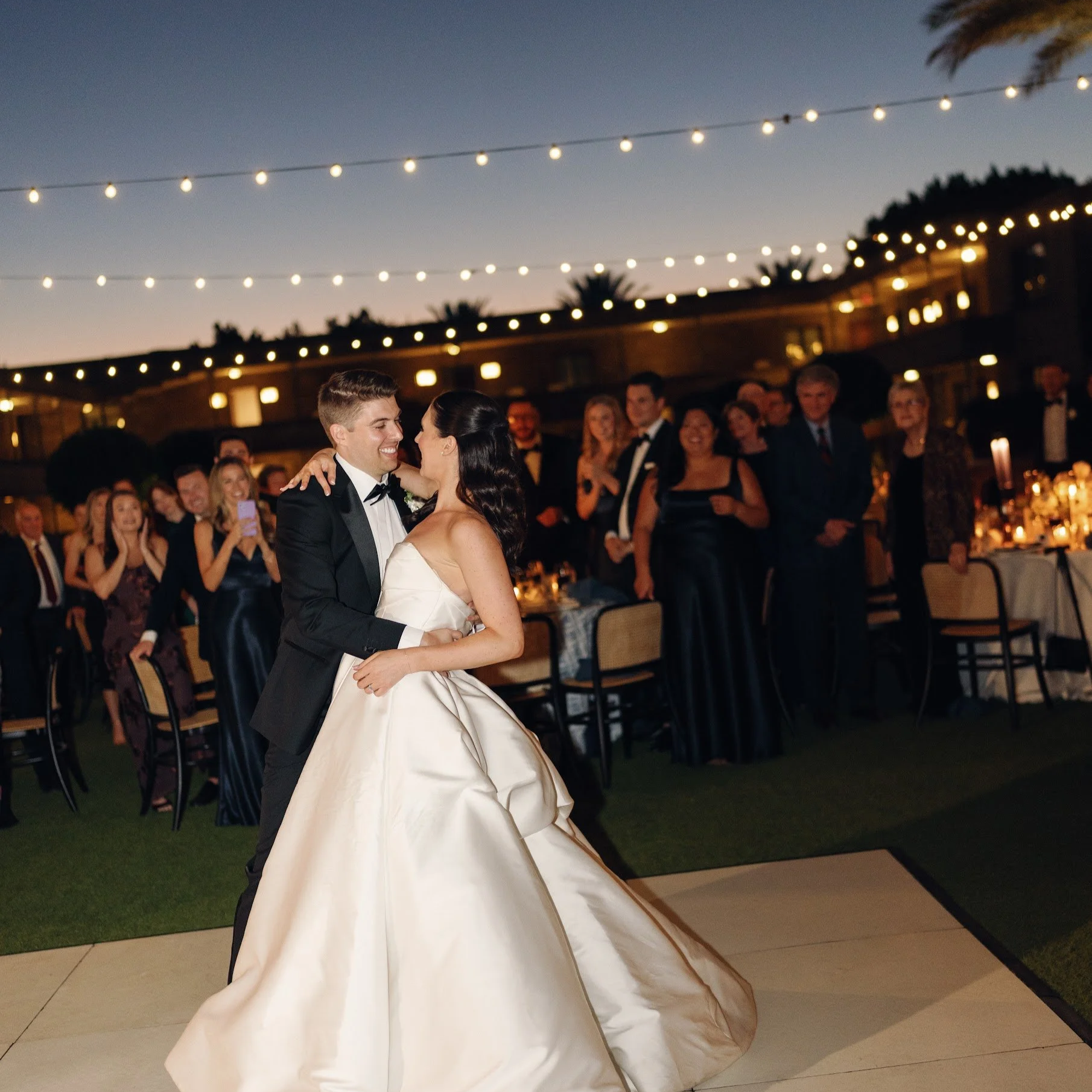 Decorated outdoor event space with round tables, white chairs, string lights, and patio heaters at dusk.