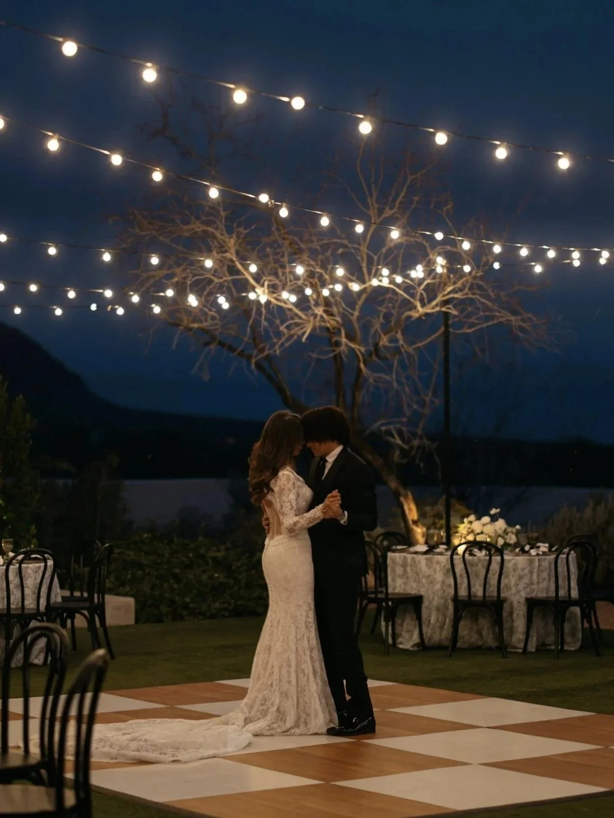Decorated outdoor event space with round tables, white chairs, string lights, and patio heaters at dusk.