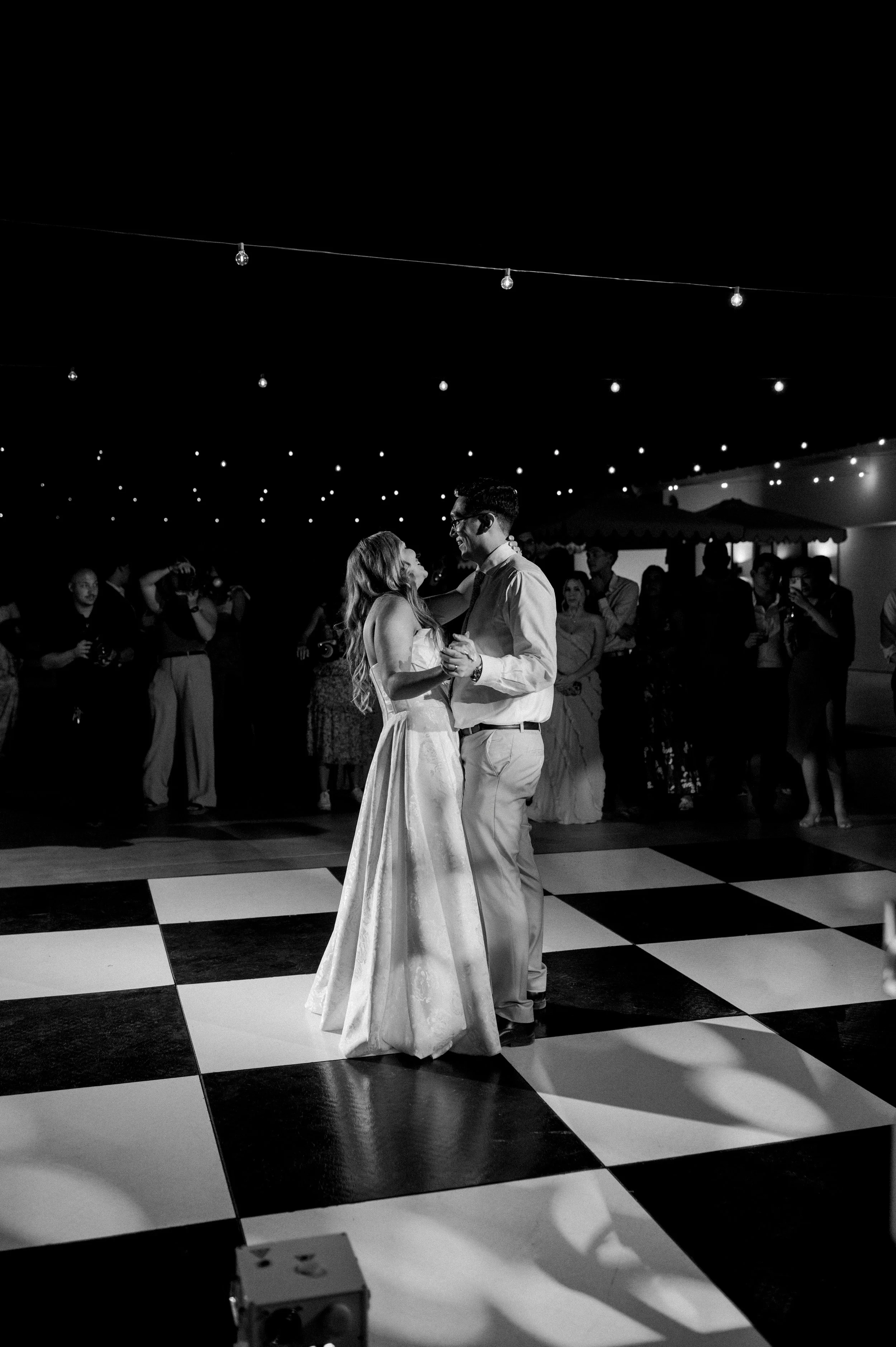 A bride and groom dancing during their wedding reception on a checkered dance floor with string lights overhead and guests watching in the background.