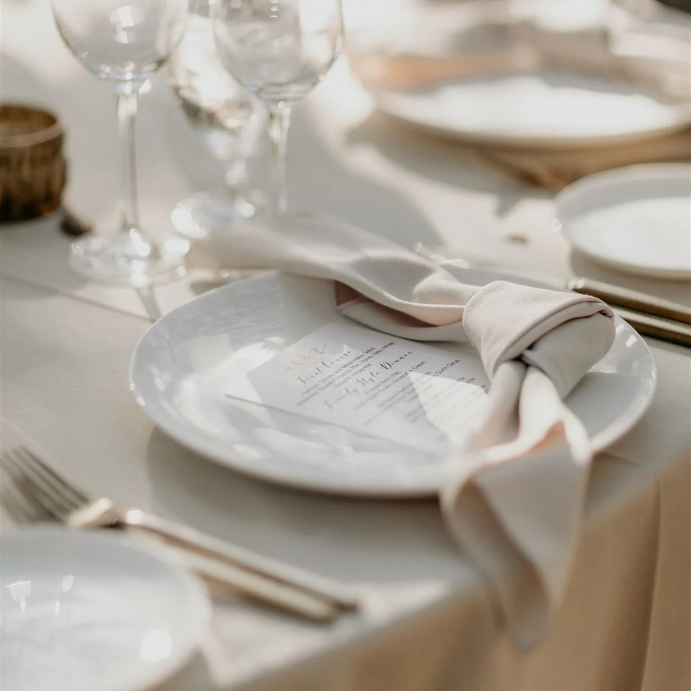 Elegant table setting with white plate, beige cloth napkin, and a menu card underneath, with wine glasses in the background.