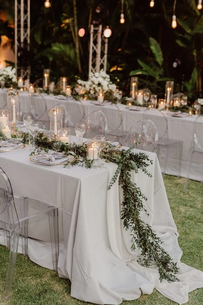 An elegantly decorated outdoor wedding reception table with white tablecloths, greenery garlands, candles, and clear chairs, set amidst a lush garden with hanging lights.