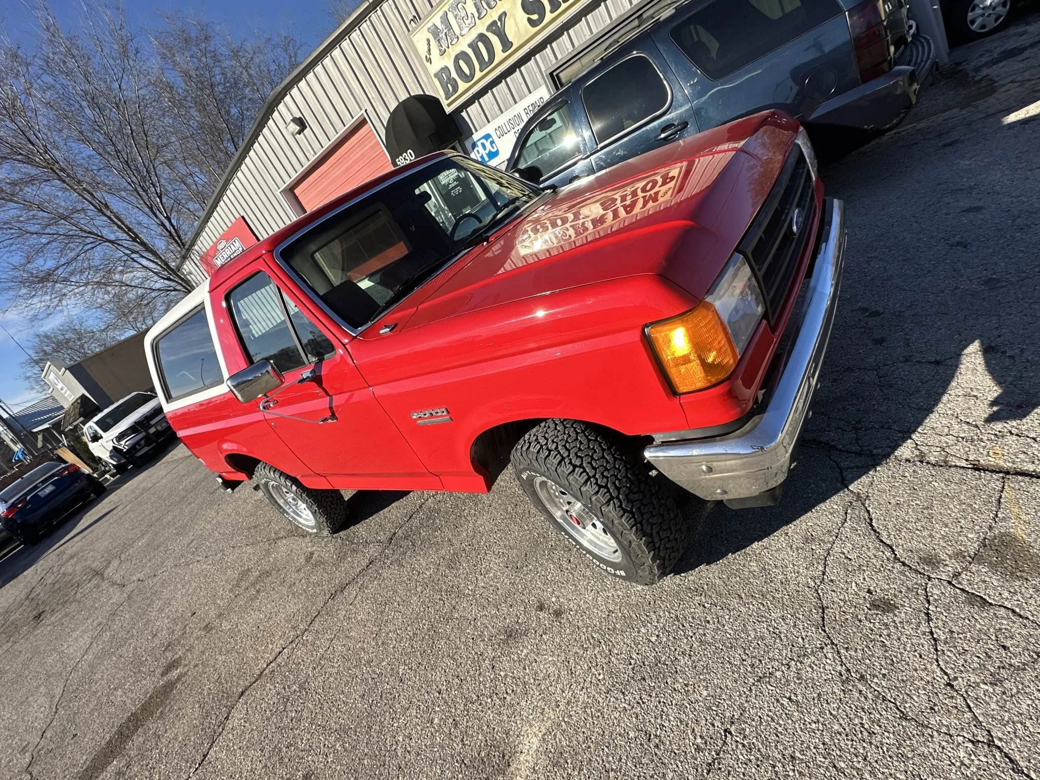 A red pickup truck parked outside a building with a sign for auto body work. The truck has black tires and appears to be in good condition.