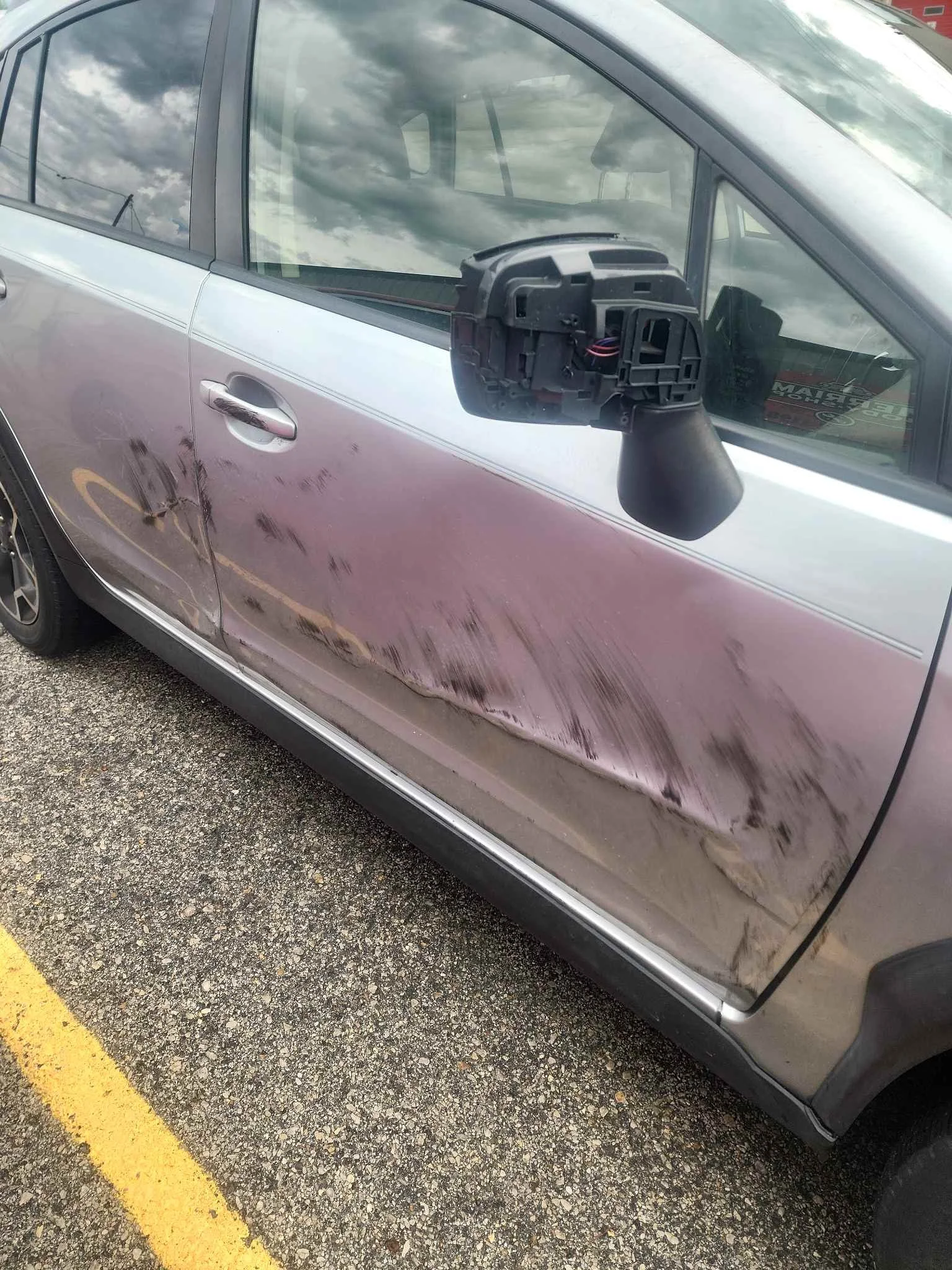 Side of a silver car with a damaged door and a side mirror that is detached, lying next to the window.