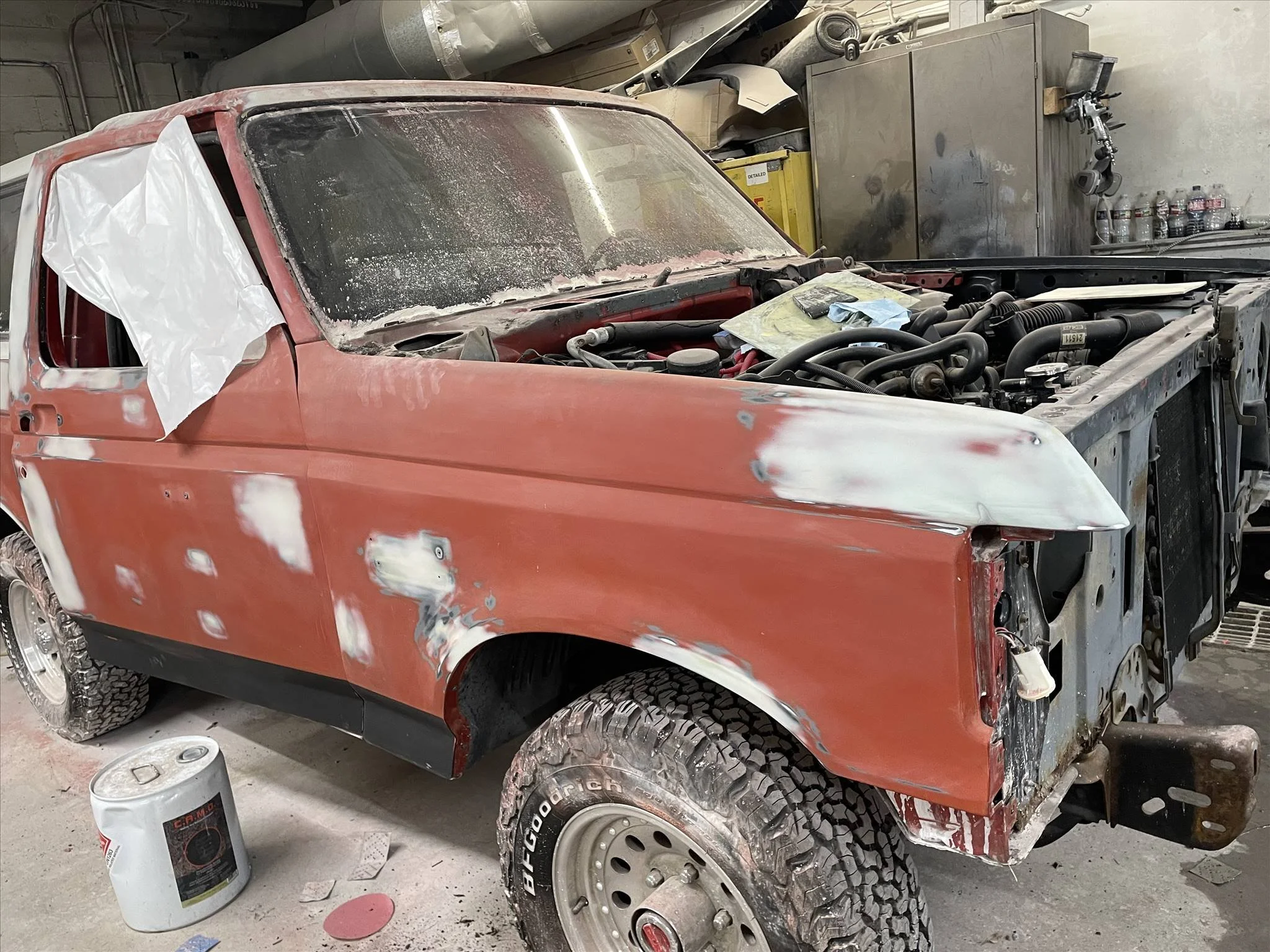 A red pickup truck undergoing restoration in a workshop. The car has primer spots, and the front hood is missing, exposing the engine. The windshield is dusty, and the side mirror and part of the door are covered with a plastic sheet. There are paint