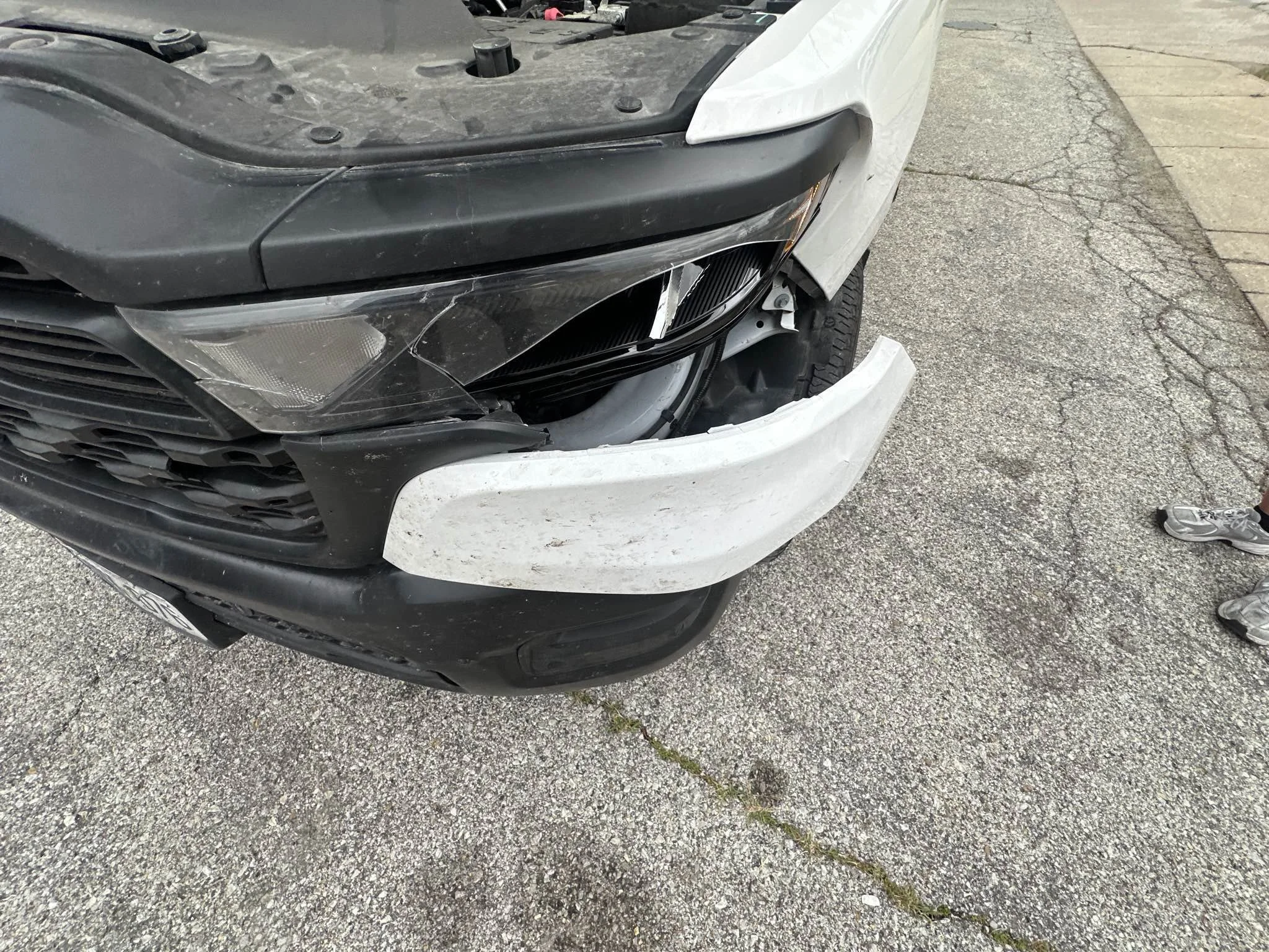 Front of a black and white car with damage to the front bumper and hood, parked on a gravel and concrete surface.