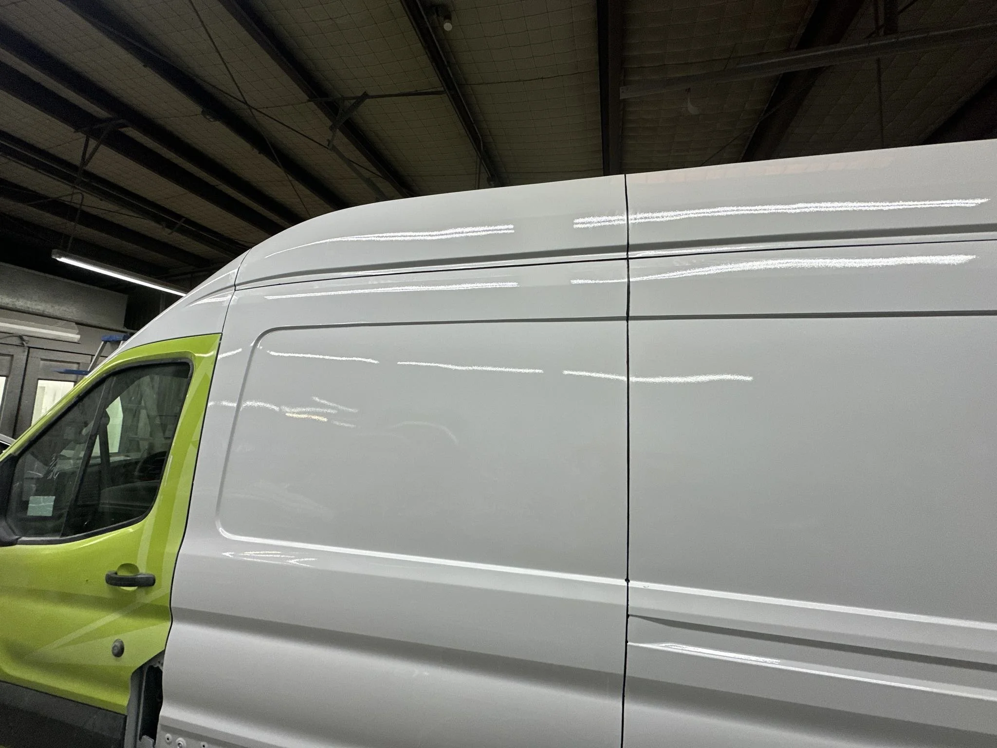 Close-up of a white commercial van with a lime green front bumper and side mirror, parked indoors with a high industrial ceiling overhead.