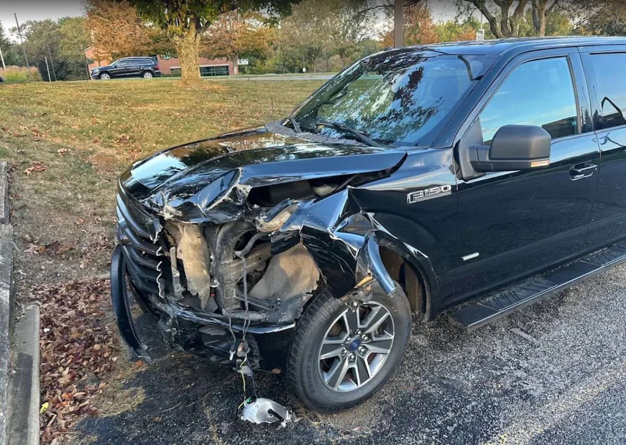 Black Ford F150 truck with front-end damage from collision, parked on the side of the road with a damaged front bumper, hood, and exposed internal components.