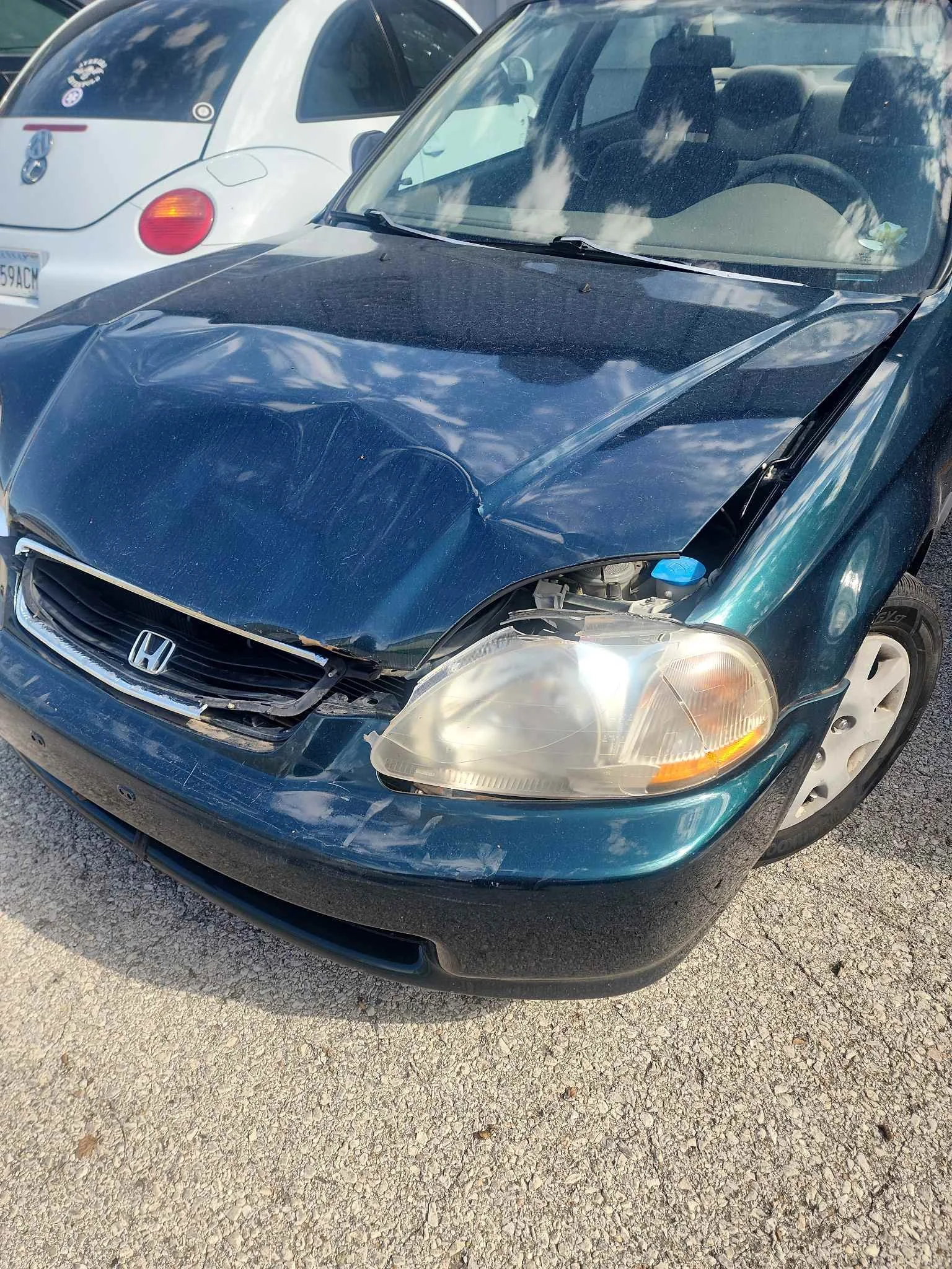 A black Honda sedan with front-end damage, including a crumpled hood and broken grille, parked on a gravel surface next to a white Volkswagen Beetle.