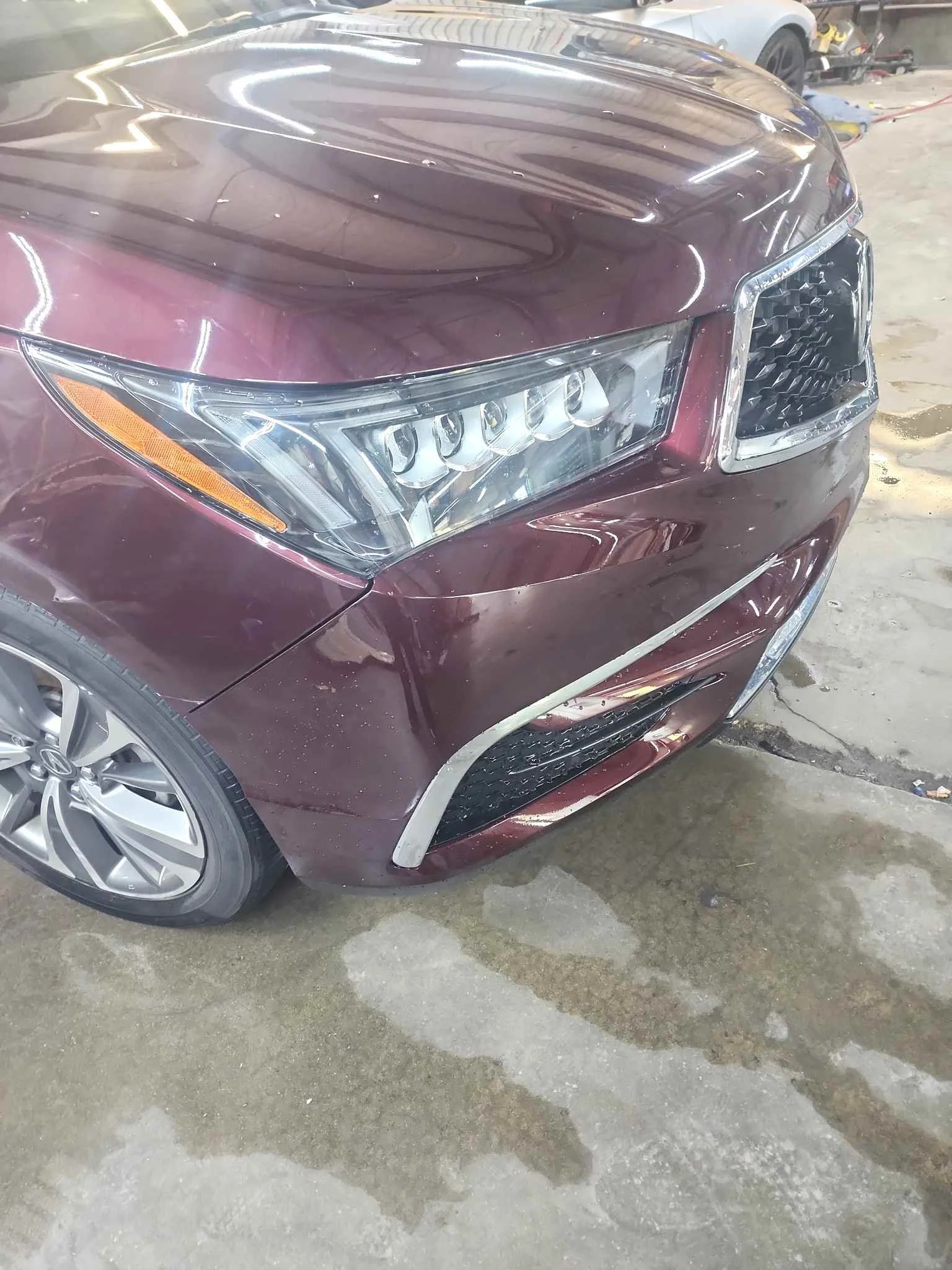 Close-up of the front of a maroon or burgundy car, showing the headlight, front grill, and part of the front wheel, in a garage or workshop with a concrete floor and other vehicles and tools in the background.
