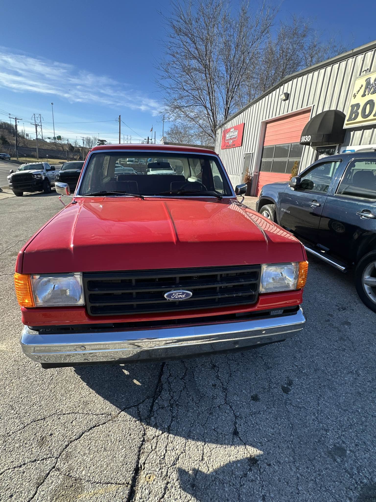 Front view of a red vintage Ford pickup truck parked in a lot with other vehicles, beside a grey industrial building with a red garage door and black awning, under a clear blue sky with leafless tree branches in the background.