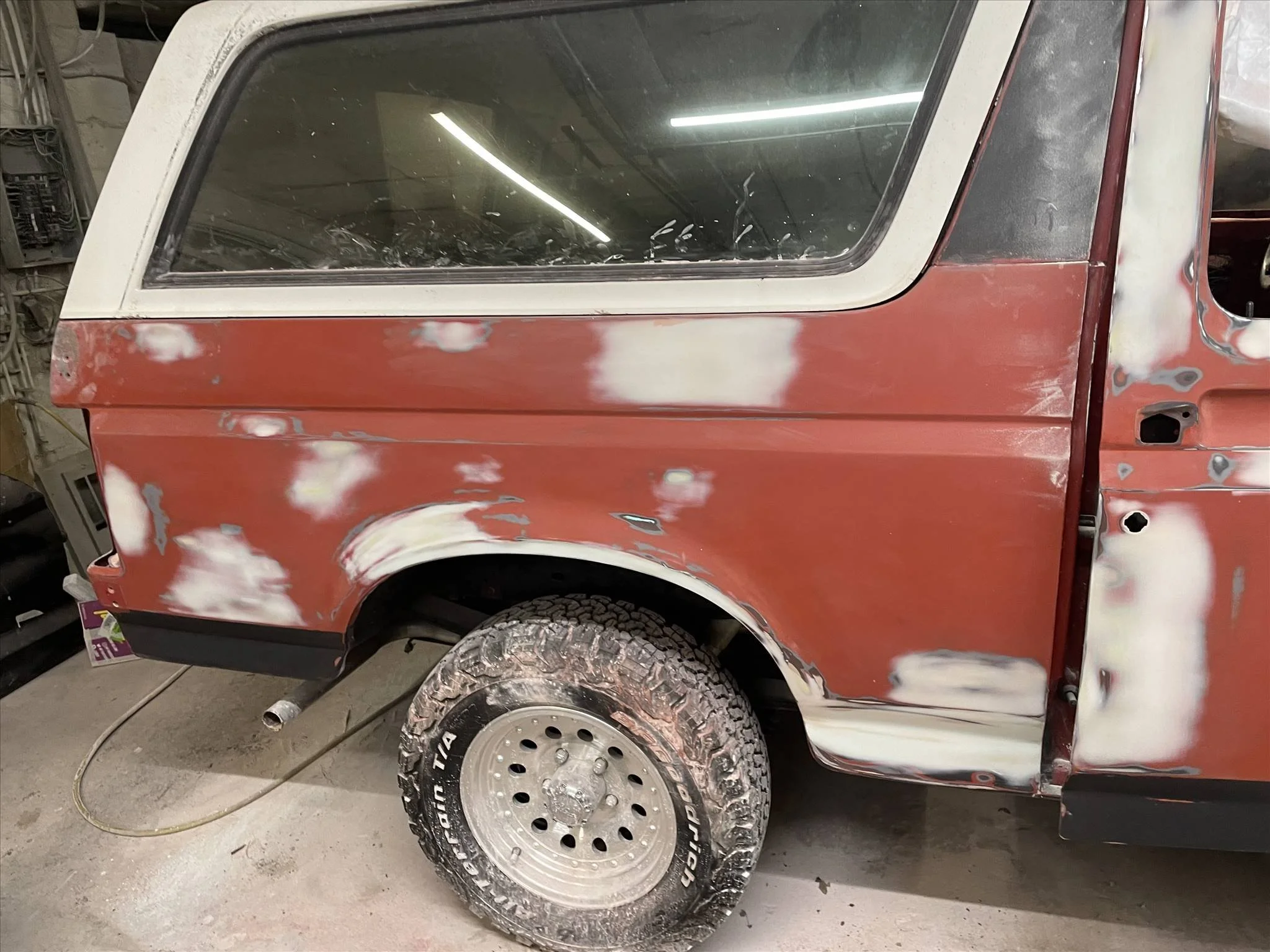 Photo of a red vehicle undergoing bodywork repair in a garage. The vehicle has patches of primer and is being prepared for repainting, with a foggy window and a rear wheel with a new tire.