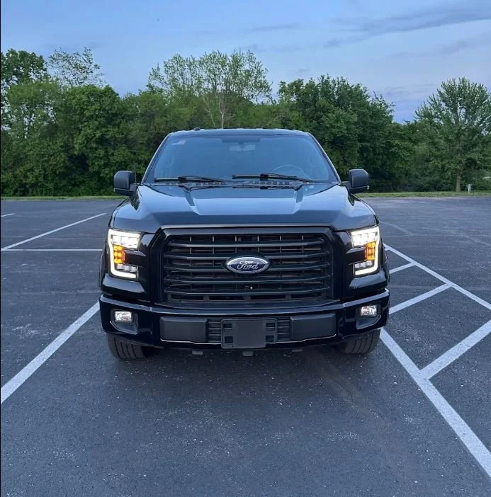 Front view of a black Ford pickup truck parked in a nearly empty parking lot with trees and a cloudy sky in the background.