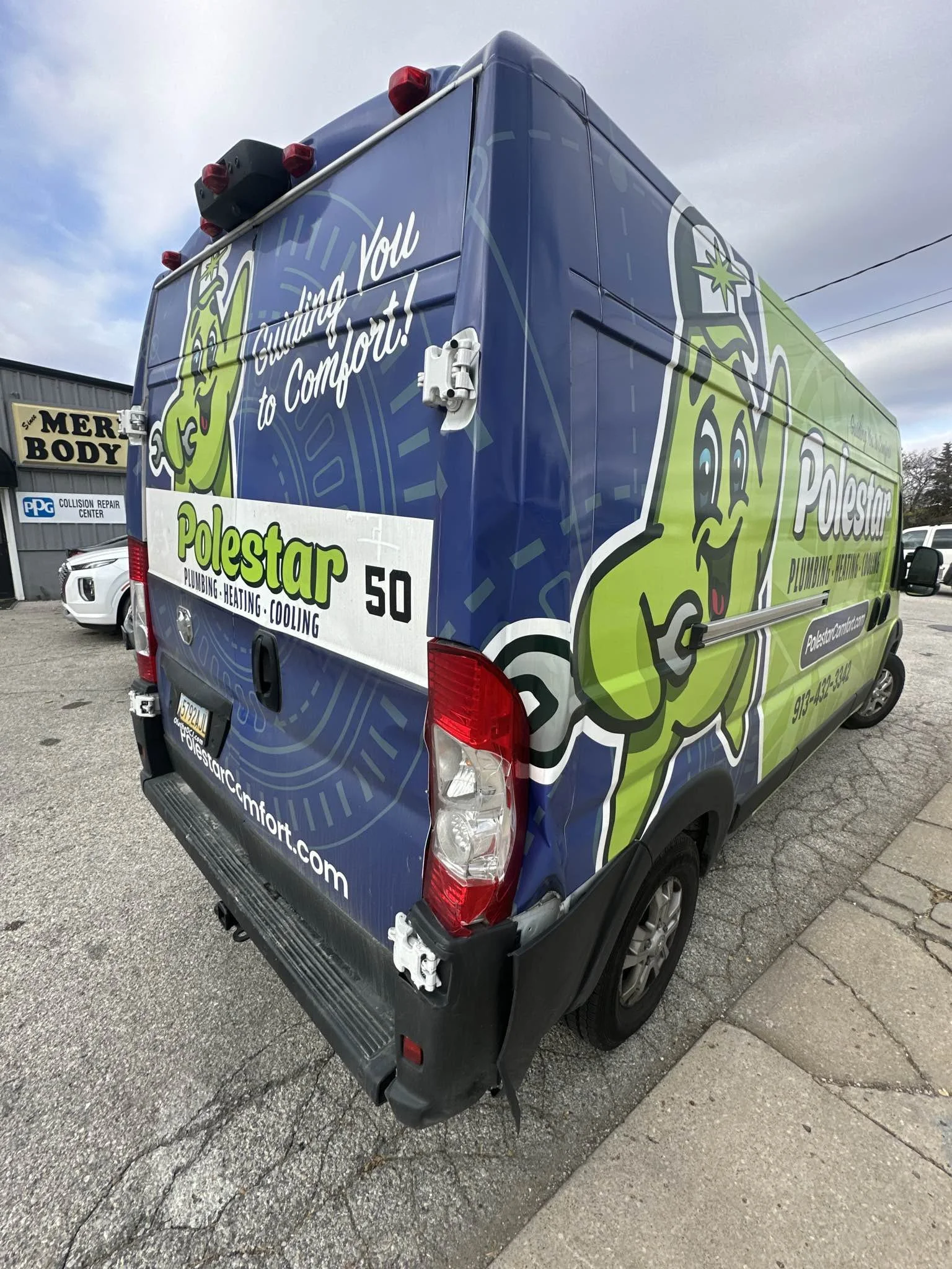 A delivery van with colorful graphics featuring a cheerful cartoon plumber character, advertising Polestar Plumbing Heating Cooling, parked on a street near a building with a sign for a collision repair center.