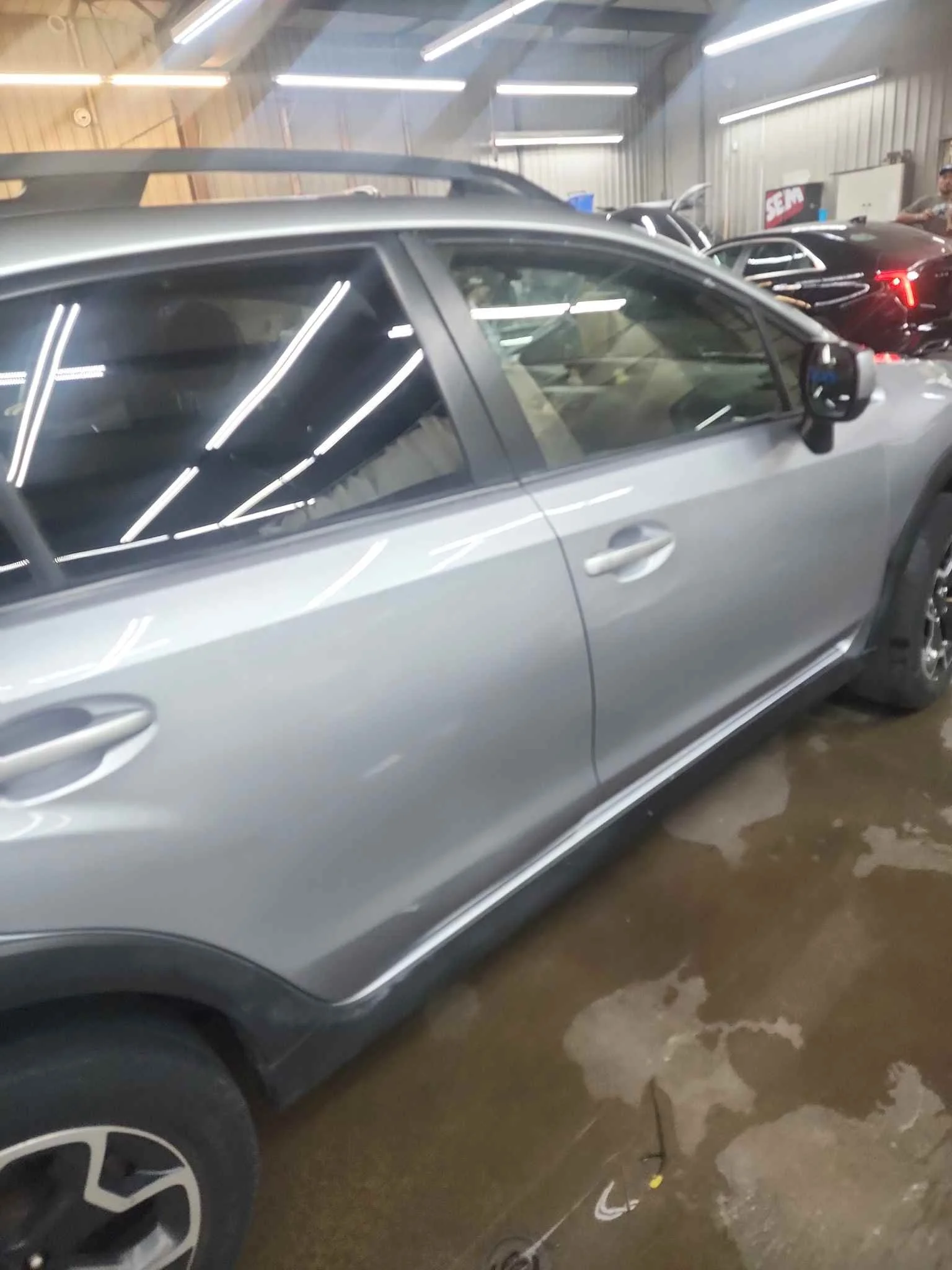Gray sedan in an indoor garage among other cars with open doors, fluorescent lighting, and visible concrete floor.