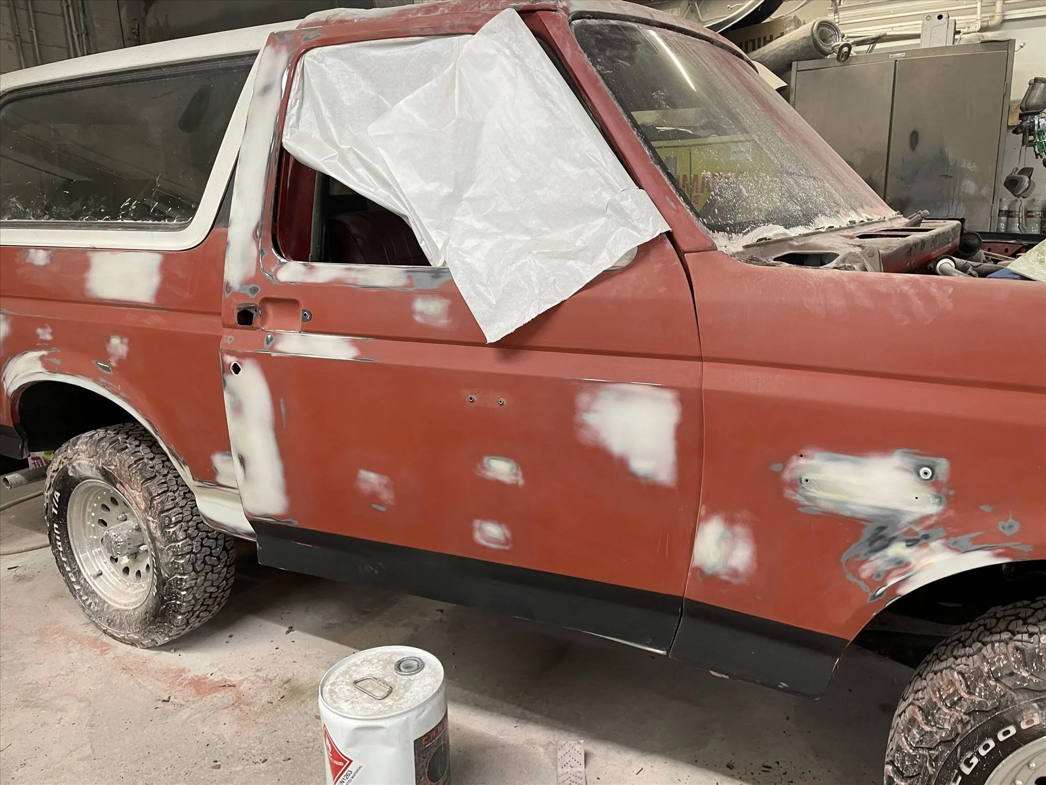 A red truck undergoing bodywork repairs in a garage, with patches of primer and sanded areas visible, a white sheet covering part of the driver side window, and typical workshop tools and containers around.