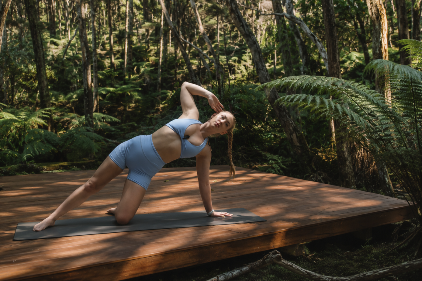A woman with long blonde hair in a black sports bra and black shorts standing outdoors with her arms wide open, enjoying sunshine in a wooded area.