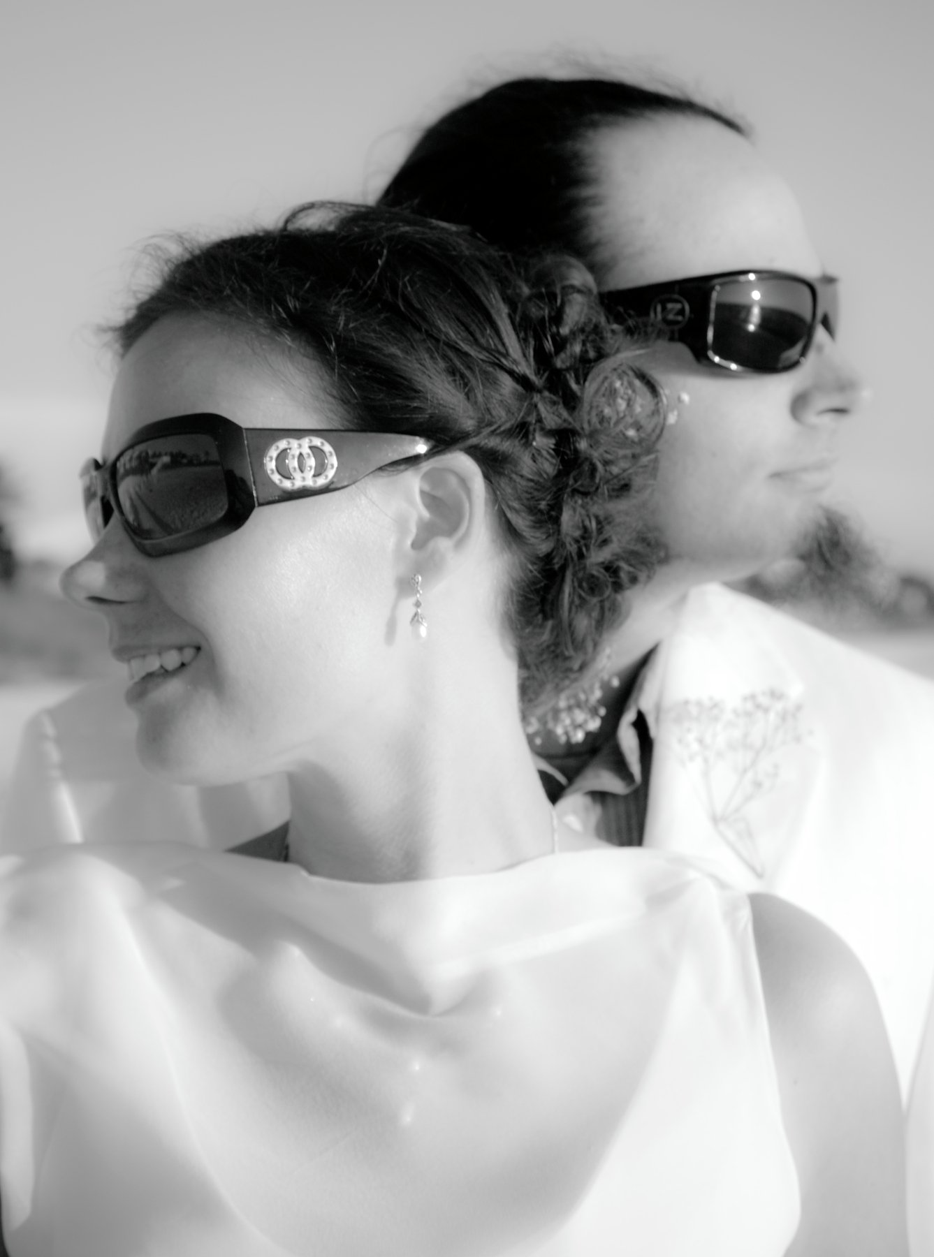 Black and white photo of a smiling woman and man wearing sunglasses, facing opposite directions, outdoors.