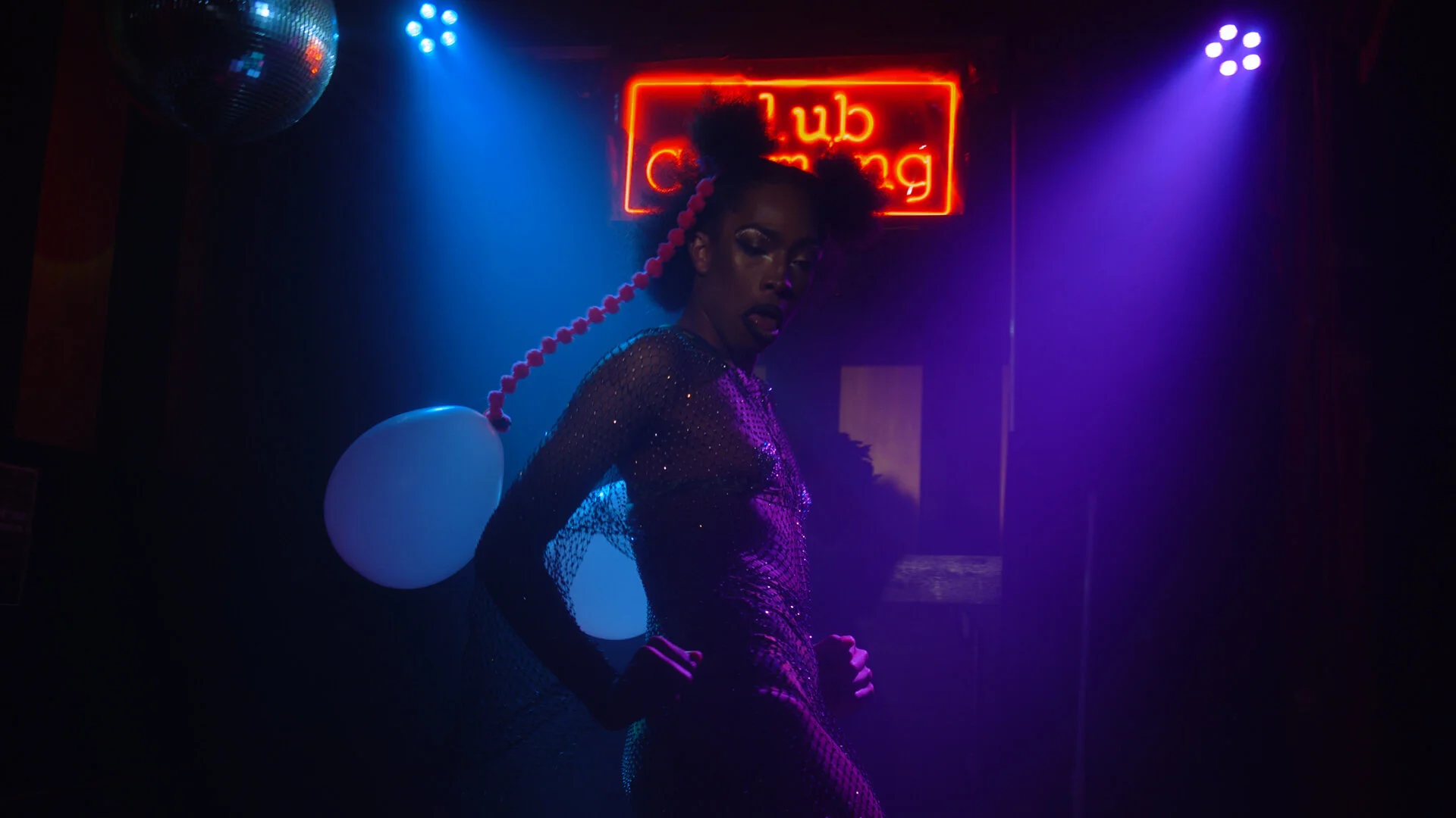 A person with curly hair styled in puffs, wearing a shiny, sequined dress, standing in front of a neon sign that reads 'club coming,' with purple and blue lighting creating a moody atmosphere.